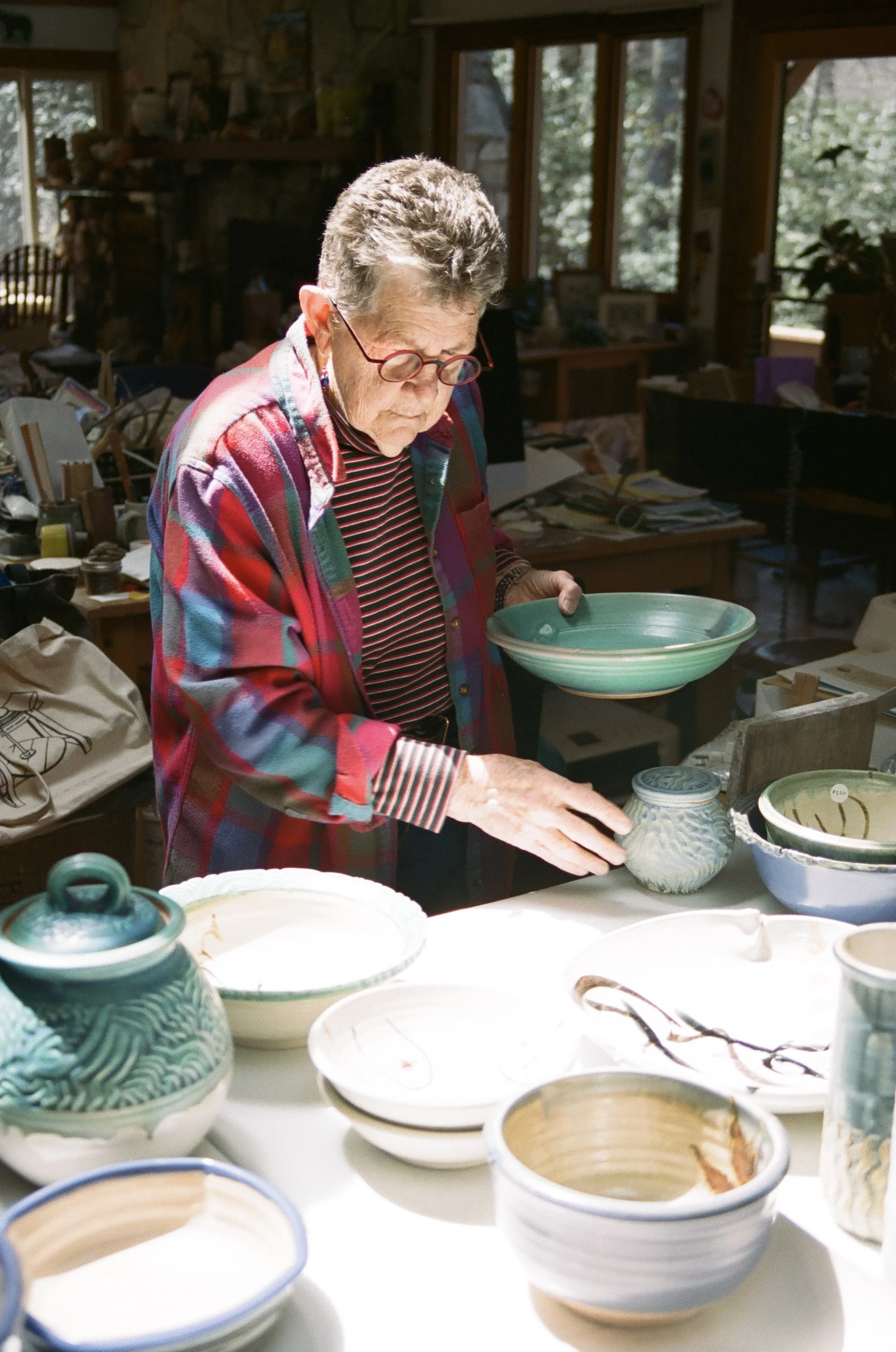 Cynthia Bringle, a professional potter, wearing glasses and a colorful, striped jacket is examining ceramic bowls and pottery on a table in a pottery studio. The studio has large windows with natural light, a stone fireplace, and various art supplies