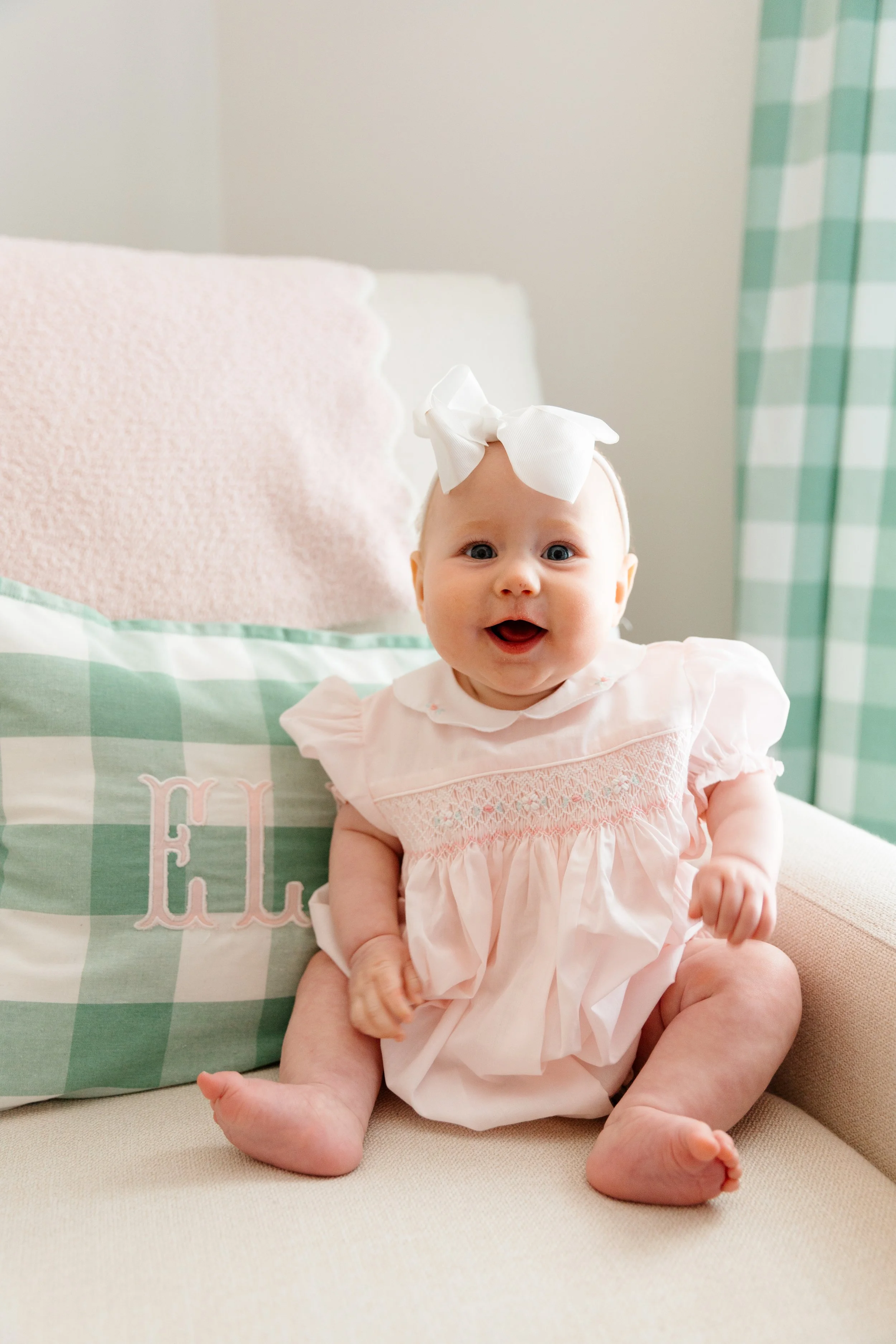Smiling baby girl Ellie with blue eyes, wearing a light pink dress with eyelet detailing and a large white bow headband, in a room with pastel-colored decor.