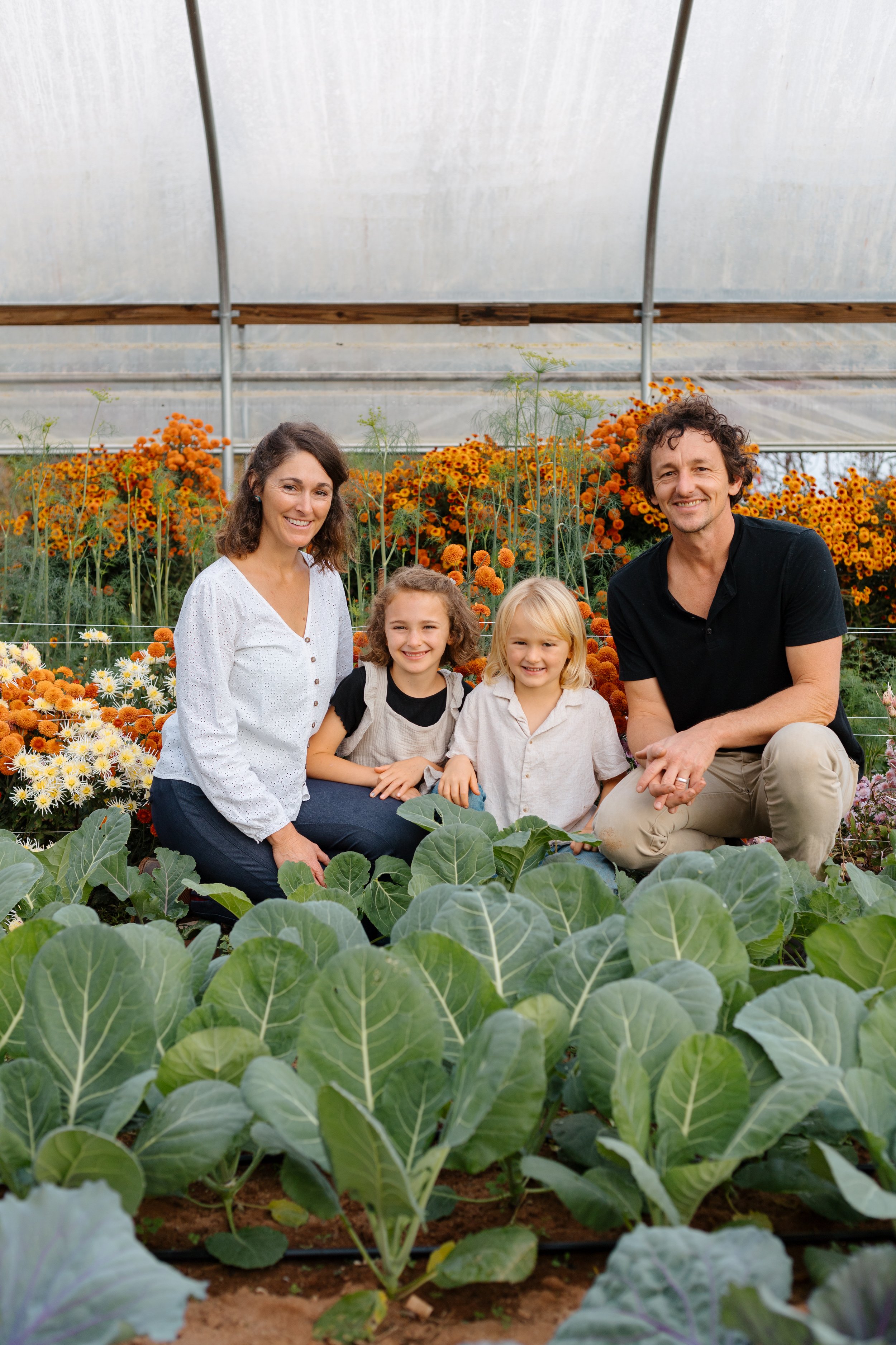 Small family farm owners, two adults and two children, kneeling among rows of leafy green plants and surrounded by orange and yellow flowers, inside a greenhouse. Pine Mountain, Georgia. 