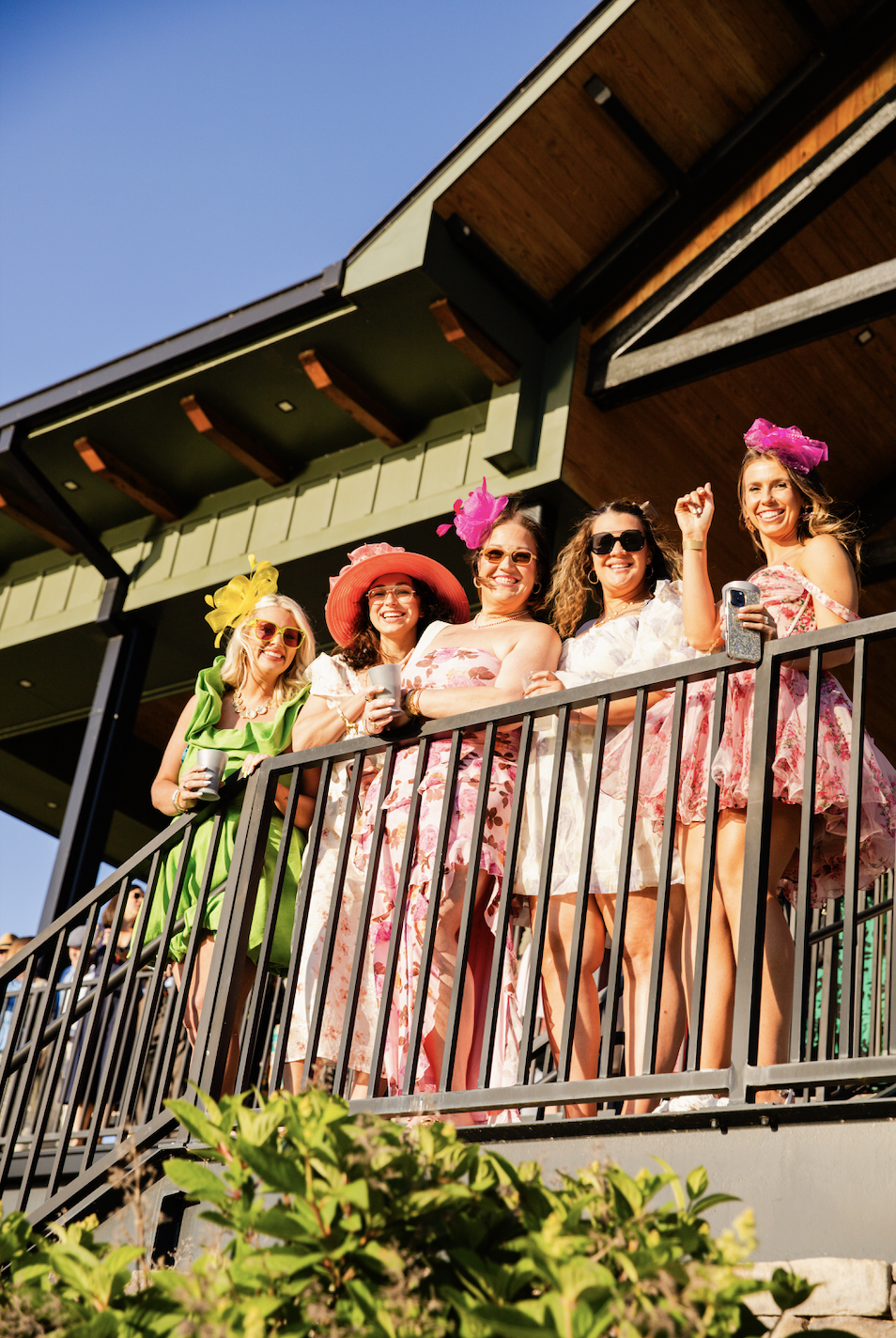 Group of five women in colorful floral dresses and large hats standing on a balcony, smiling and enjoying the sunny day. Derby party event photos. 