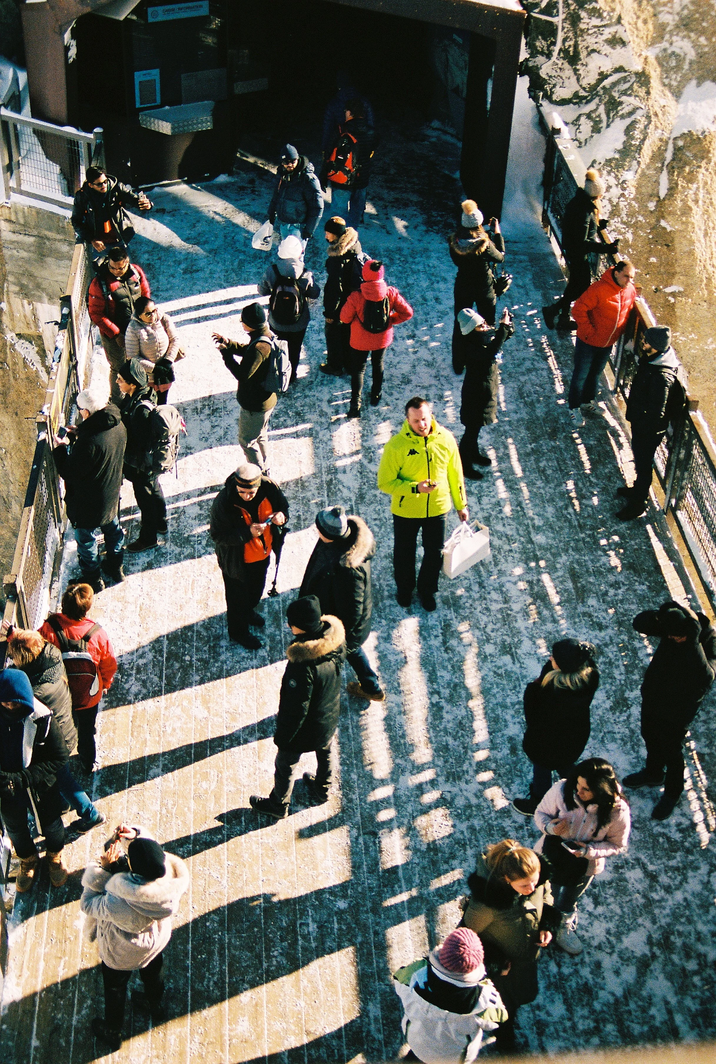 A group of people standing and walking on a snowy outdoor pathway, with some dressed in winter clothing and others taking photos, sunlight casting long shadows in Chamonix, France. 
