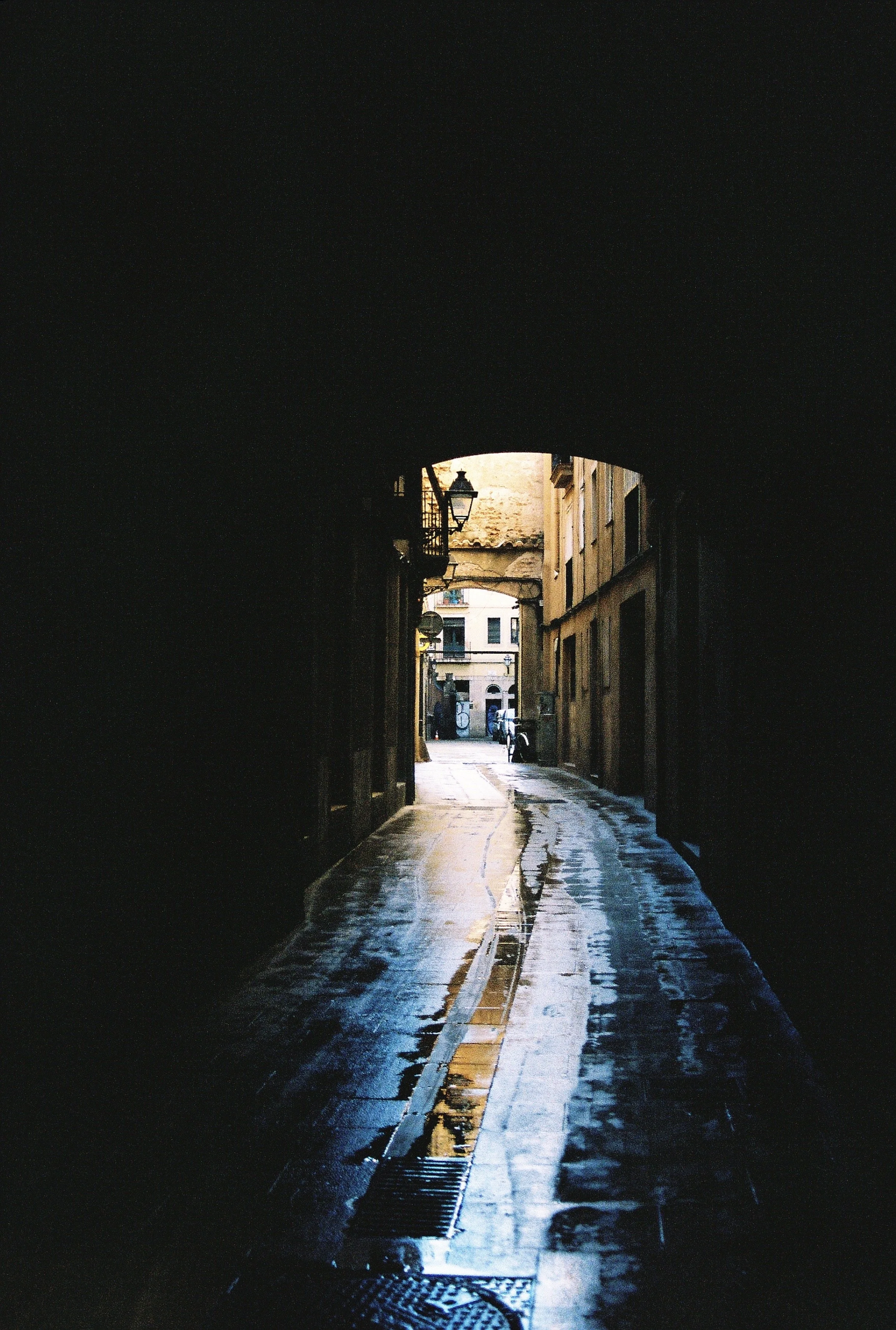 A narrow alleyway with wet pavement and puddles, leading to a sunlit street with buildings and parked cars, viewed from under an arch or tunnel.