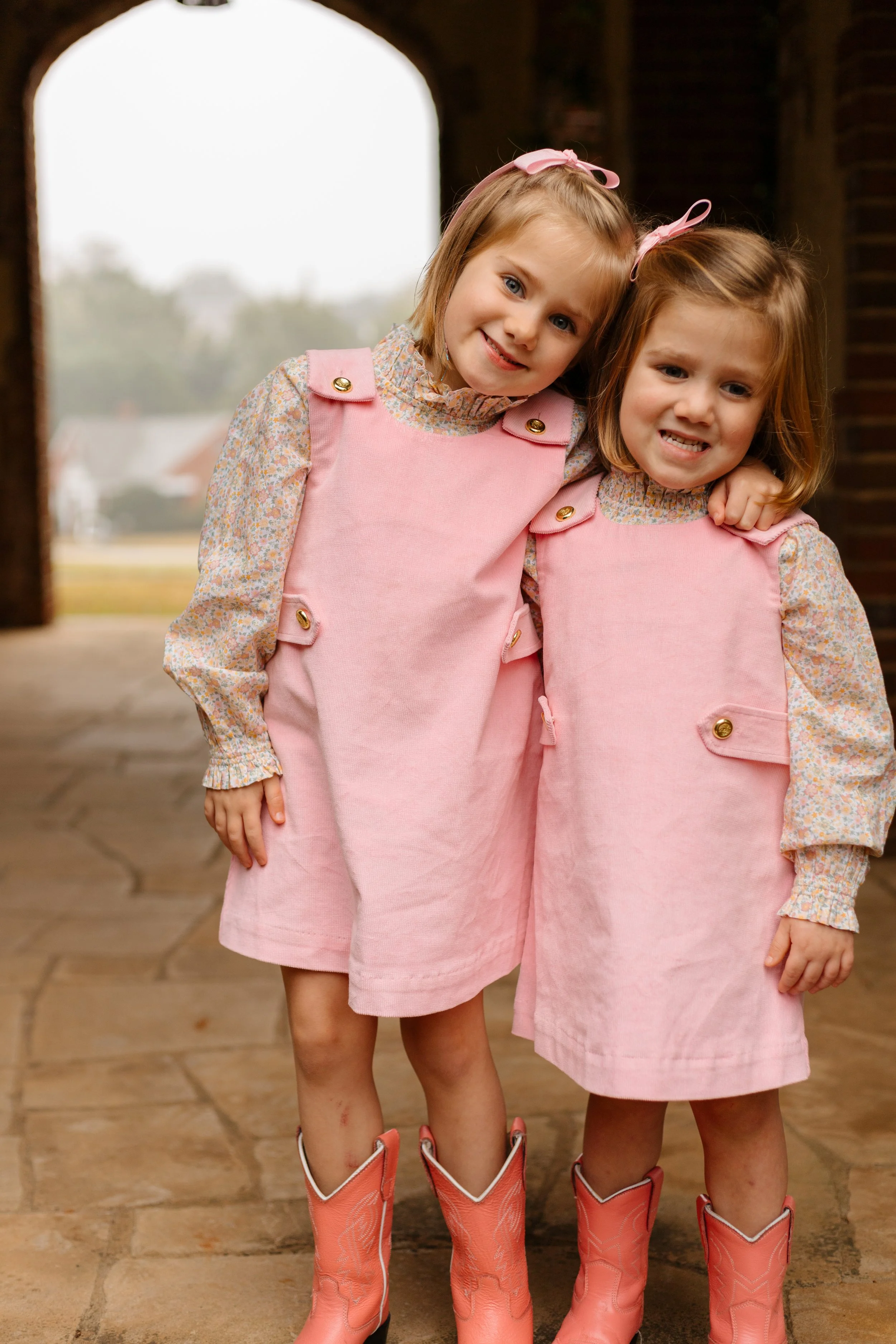 Two young girl sisters with blonde and red hair, wearing pink dresses and pink cowboy boots, standing close together outdoors under an arched structure with a blurred landscape in the background.