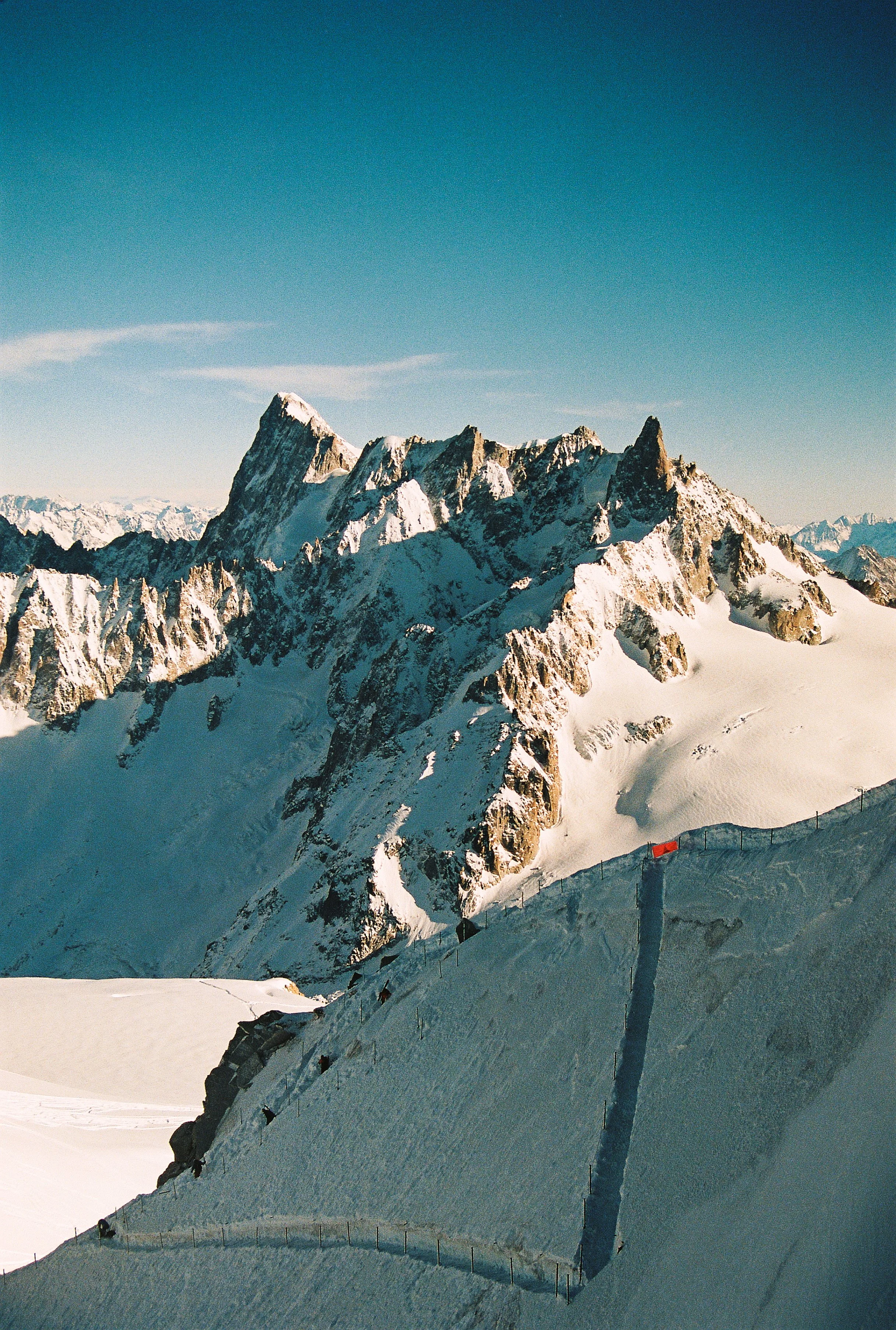 Snow-covered mountain peaks in Chamonix, France under a clear blue sky, with a snow-lined gravel path and fencing in the foreground.