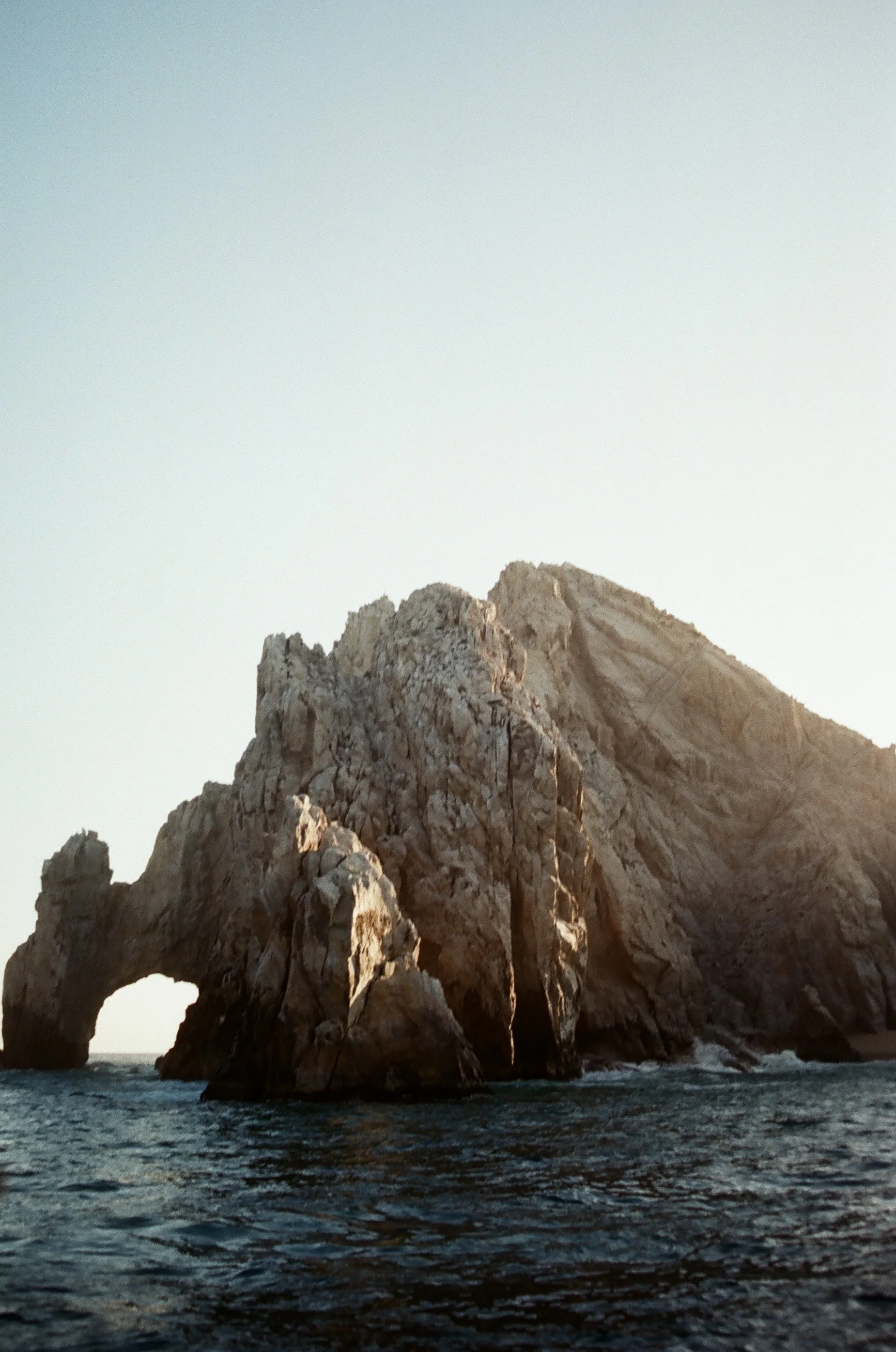 A large natural rock formation or sea stack in Cabo, Mexico, with an arch at its base, rising from the ocean, taken during sunset or sunrise.