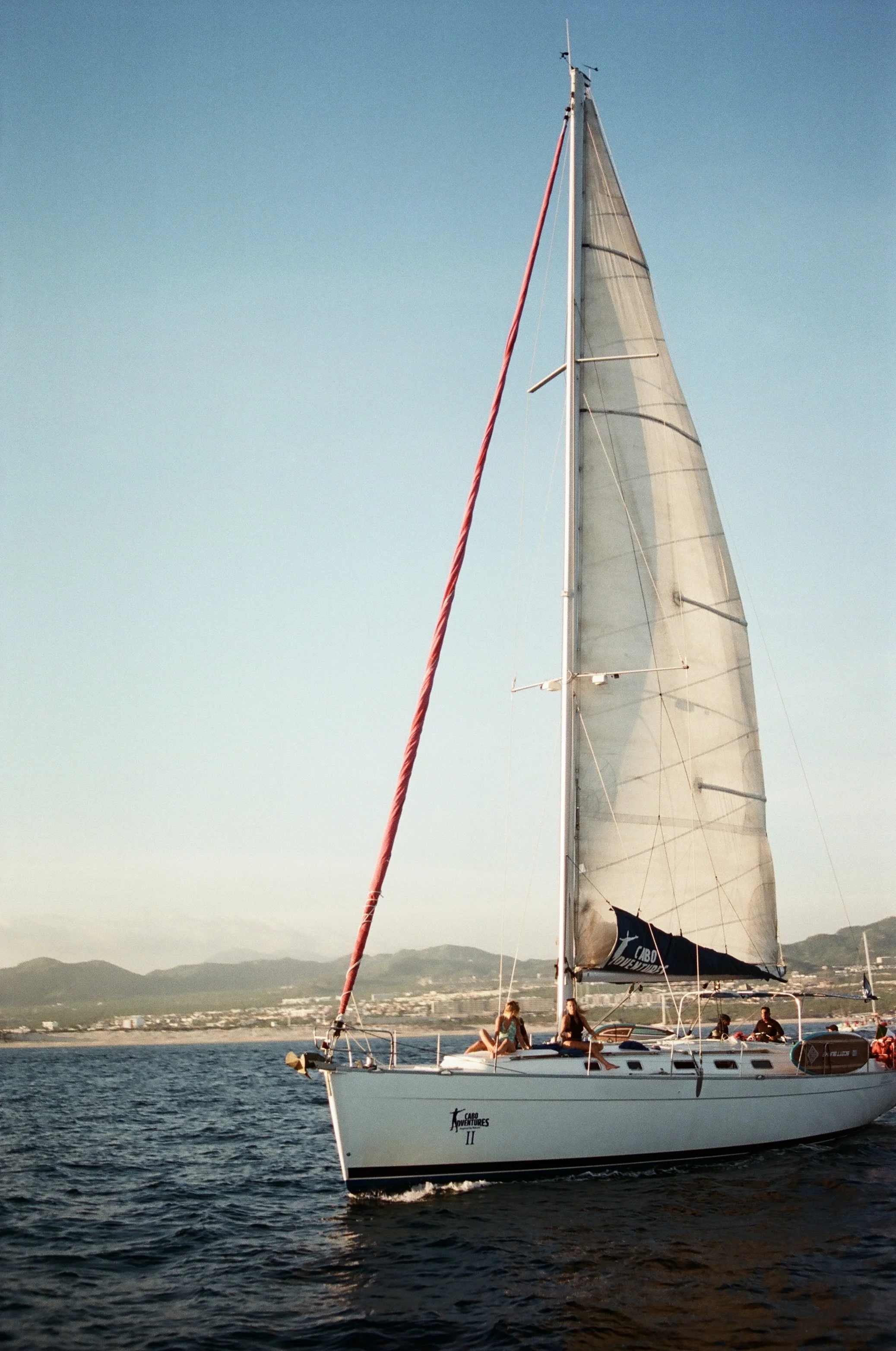 A sailboat with a white hull and a large sail on a body of water, with four people sitting on the deck, near mountains and a town in the background.