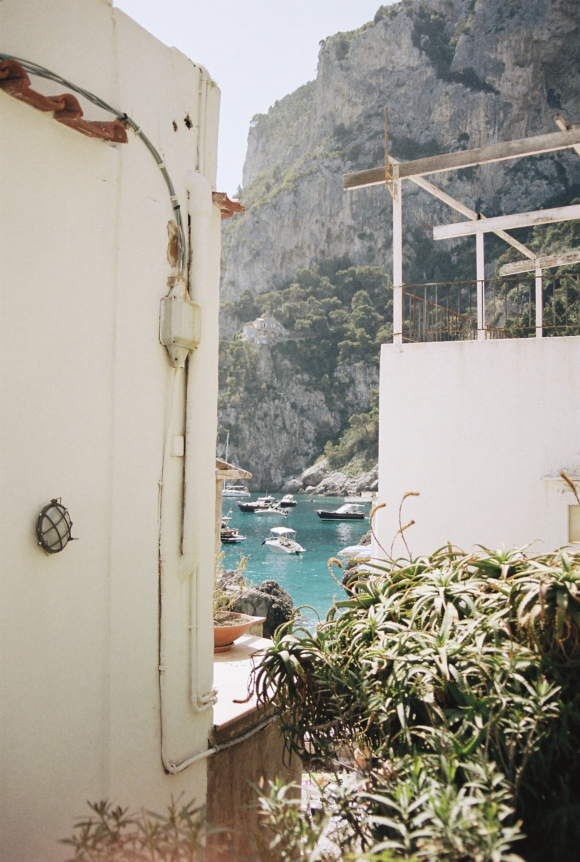 View of a rocky coastline with boats anchored in turquoise waters, seen through an open doorway with white walls, plants, and a railing in the foreground.