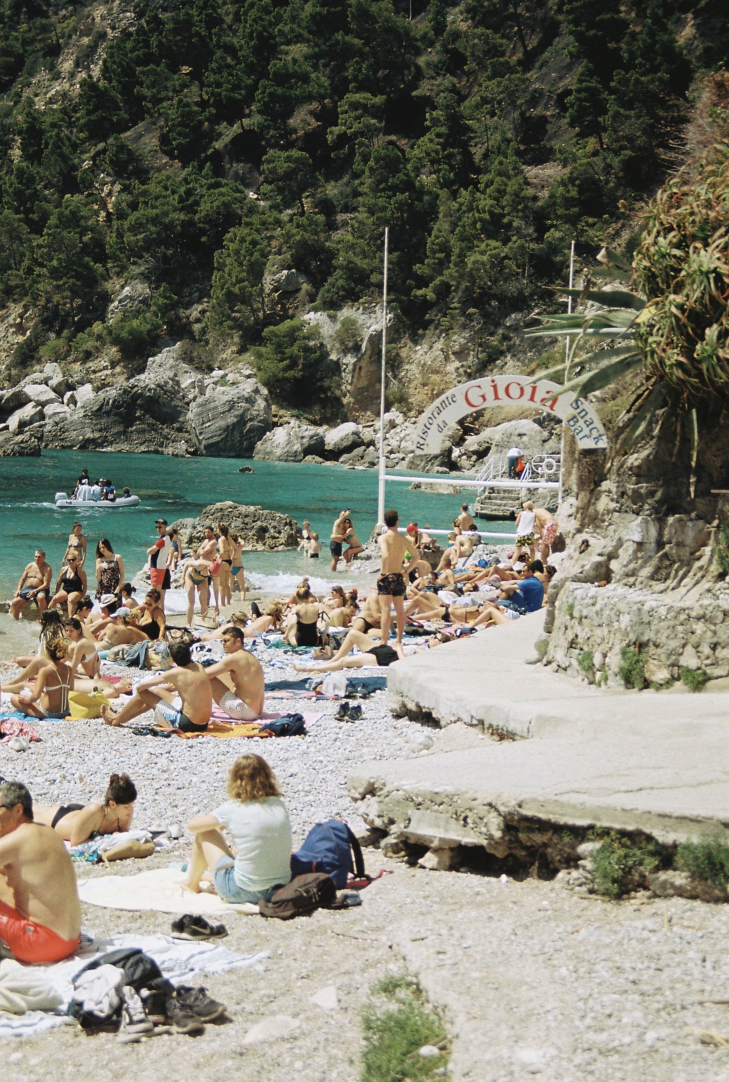 People lounging and swimming on a rocky beach with a green hillside and boats in the background in Italy. 