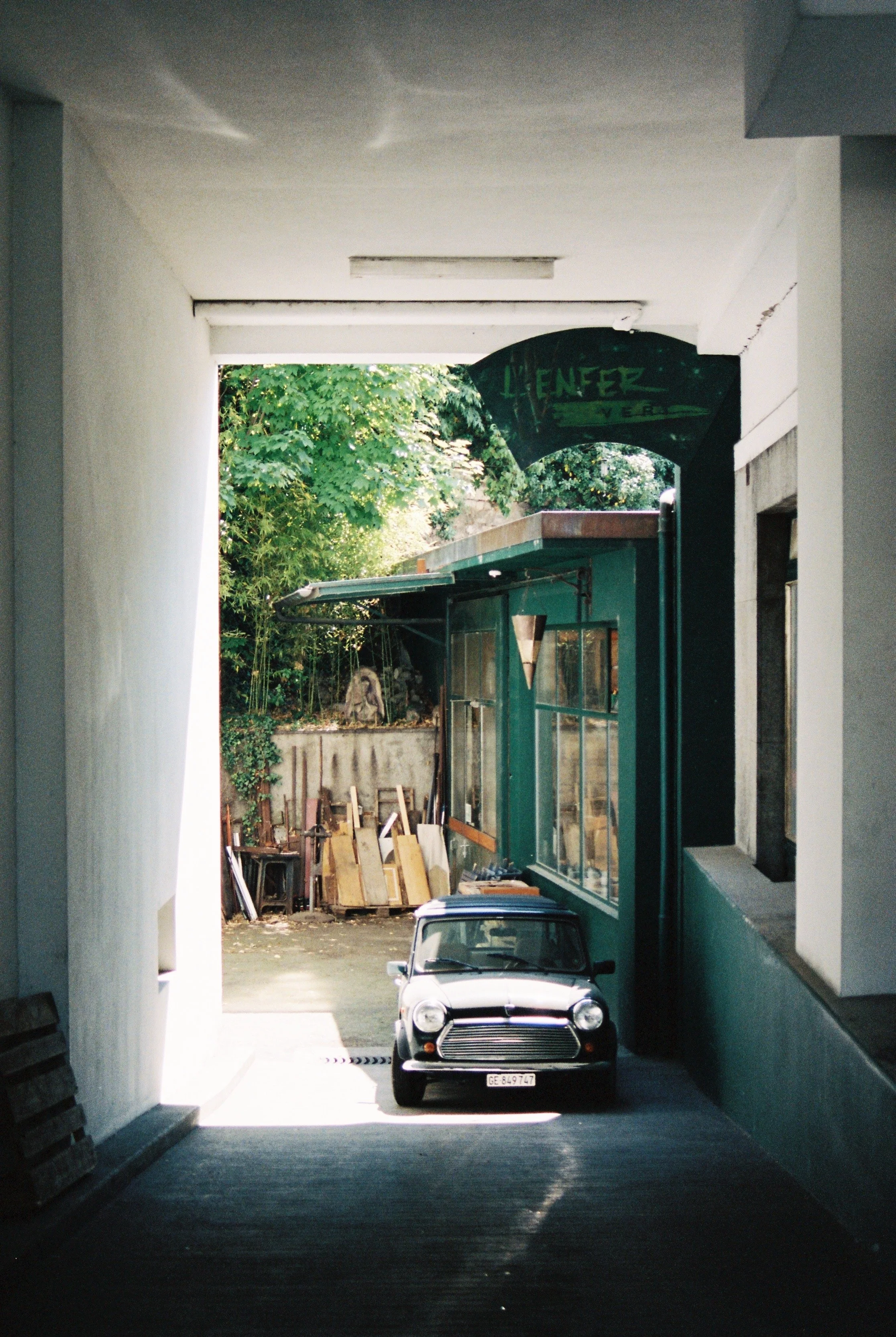 A view through a passageway showing a small vintage car parked outside on the street with a building and trees in the background in Geneva, Switzerland.