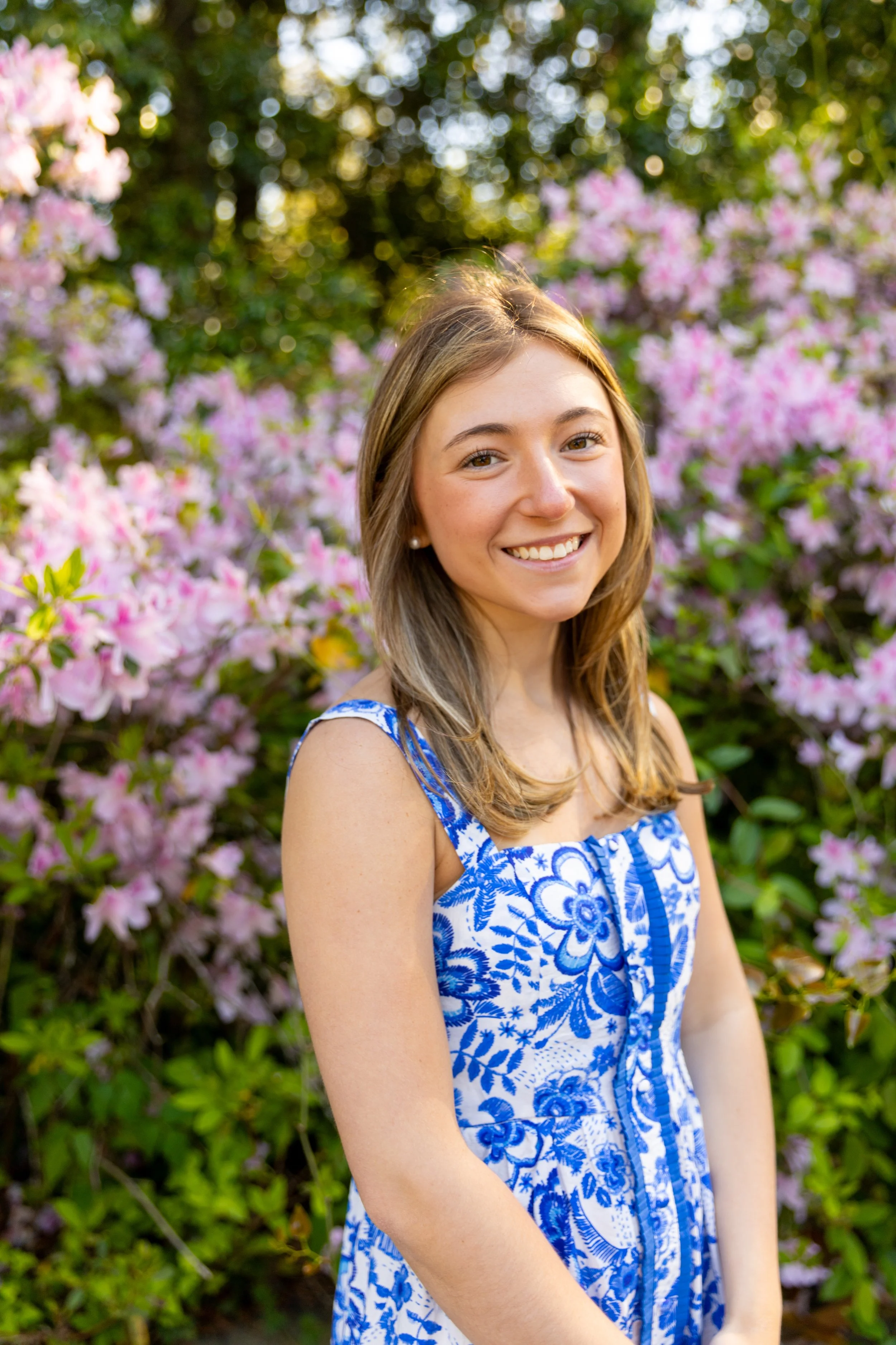 A young woman with long light brown hair, smiling, standing outdoors in front of pink and purple flowering bushes and green foliage. Senior high school photos. 