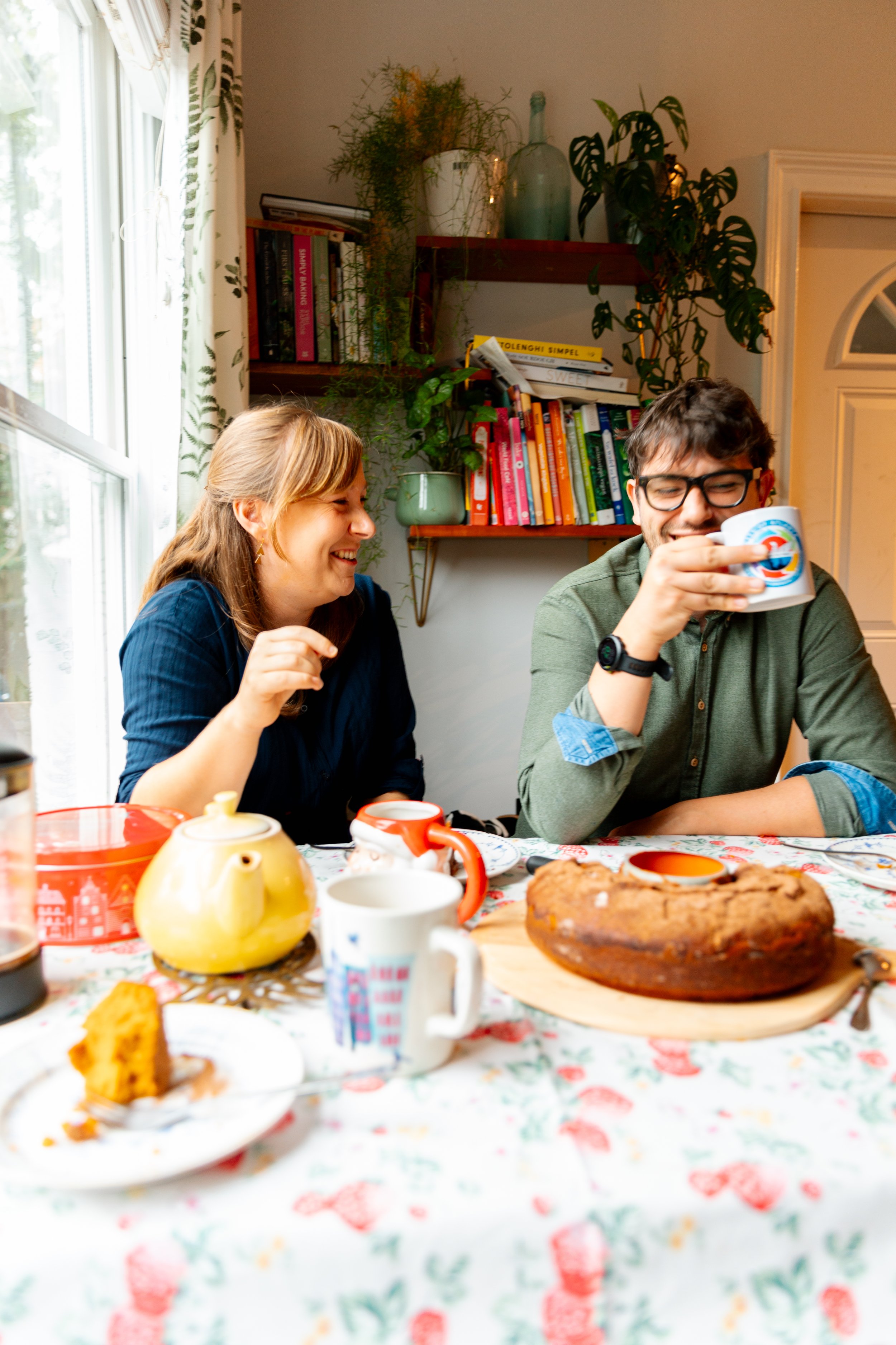 Two people sitting at a dining table with a cake, teapot, cups, and assorted breakfast food, smiling and enjoying a lively conversation in a well-lit room with bookshelves and plants.