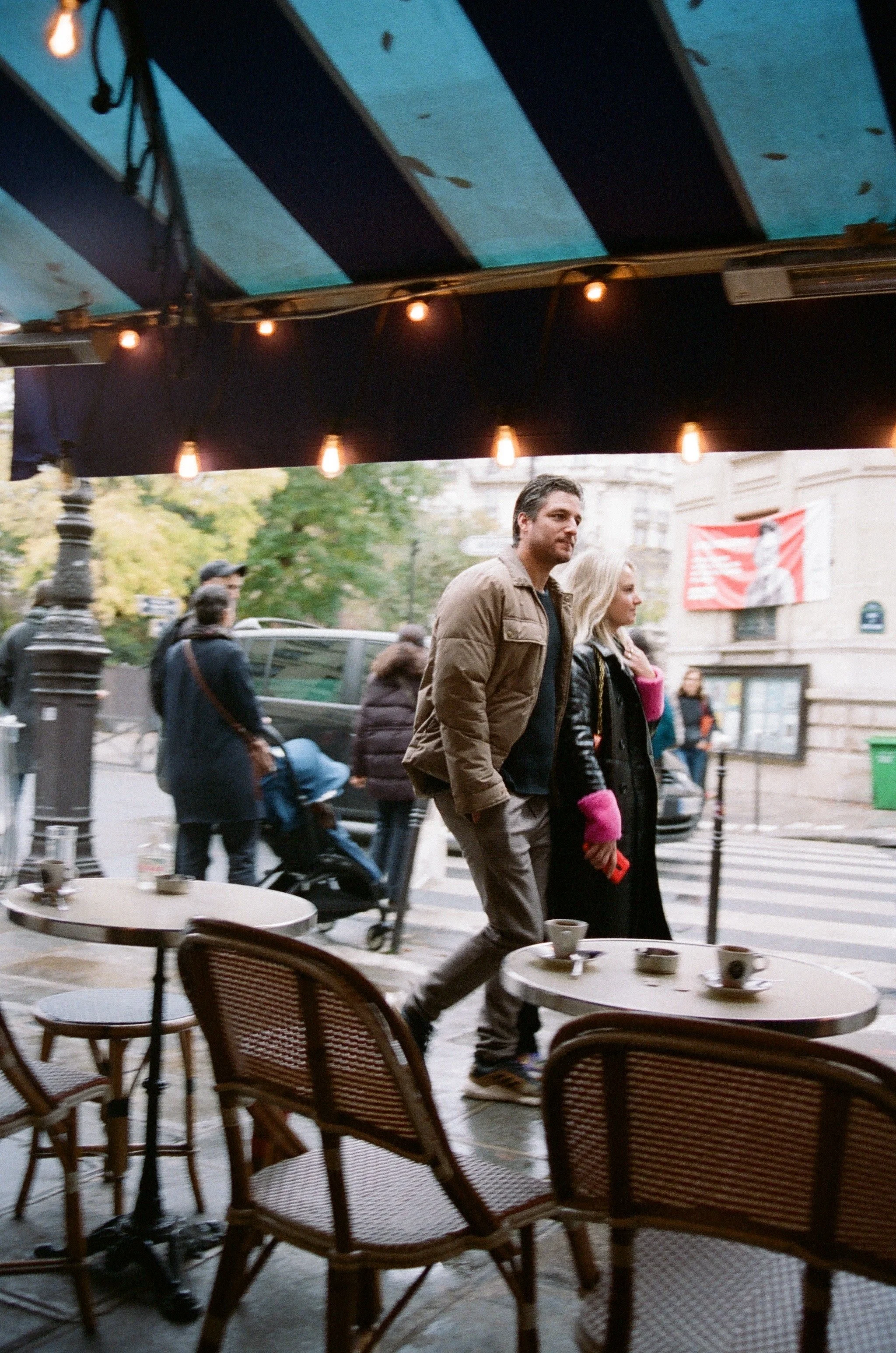 A man and a woman walking past an outdoor cafe on a city street in Paris, France, with tables and chairs, during daytime.