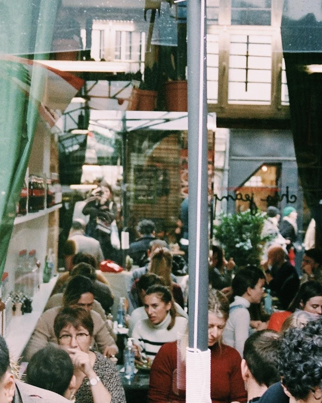 Inside a crowded café, people are sitting at tables and standing, with large windows reflecting the street outside.