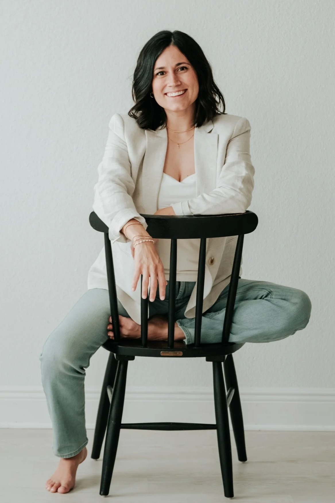 Woman sitting cross-legged on a black chair, smiling, with a white background, wearing a white blazer, white top, and gray pants.