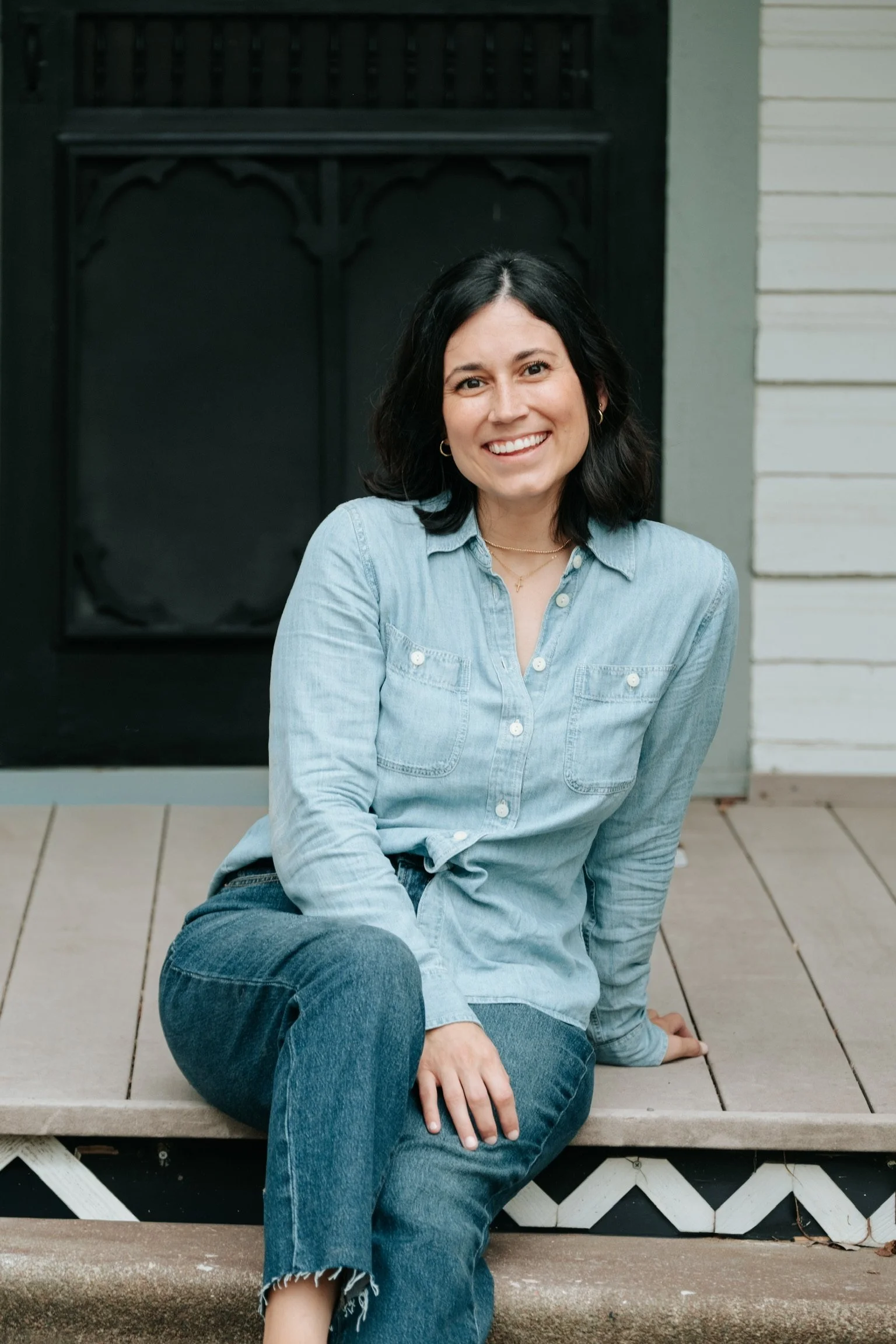 A woman with shoulder-length dark hair is sitting on a porch step, smiling at the camera. She is wearing a light blue denim shirt and jeans. The porch has a wooden lattice and a white wall with horizontal siding in the background.