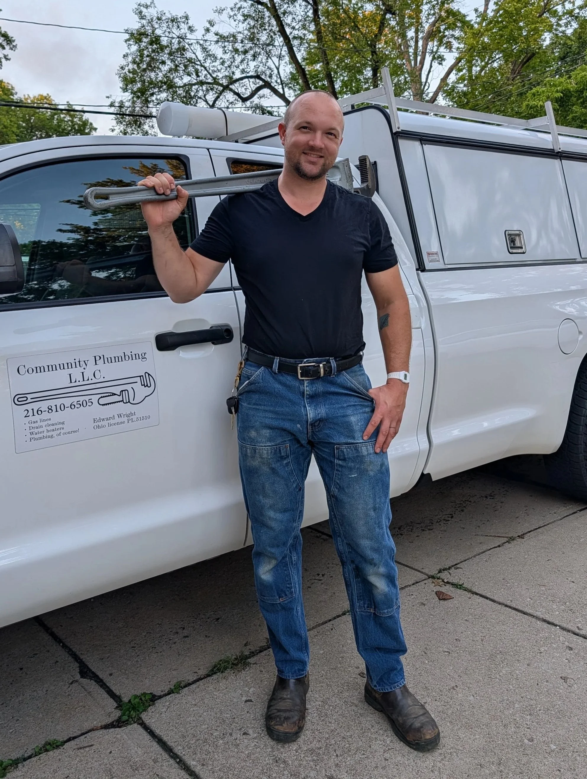 A man standing outdoors in front of a white service truck holding a large pipe wrench over his shoulder, wearing a black t-shirt, jeans, and work boots, with trees and a cloudy sky in the background.