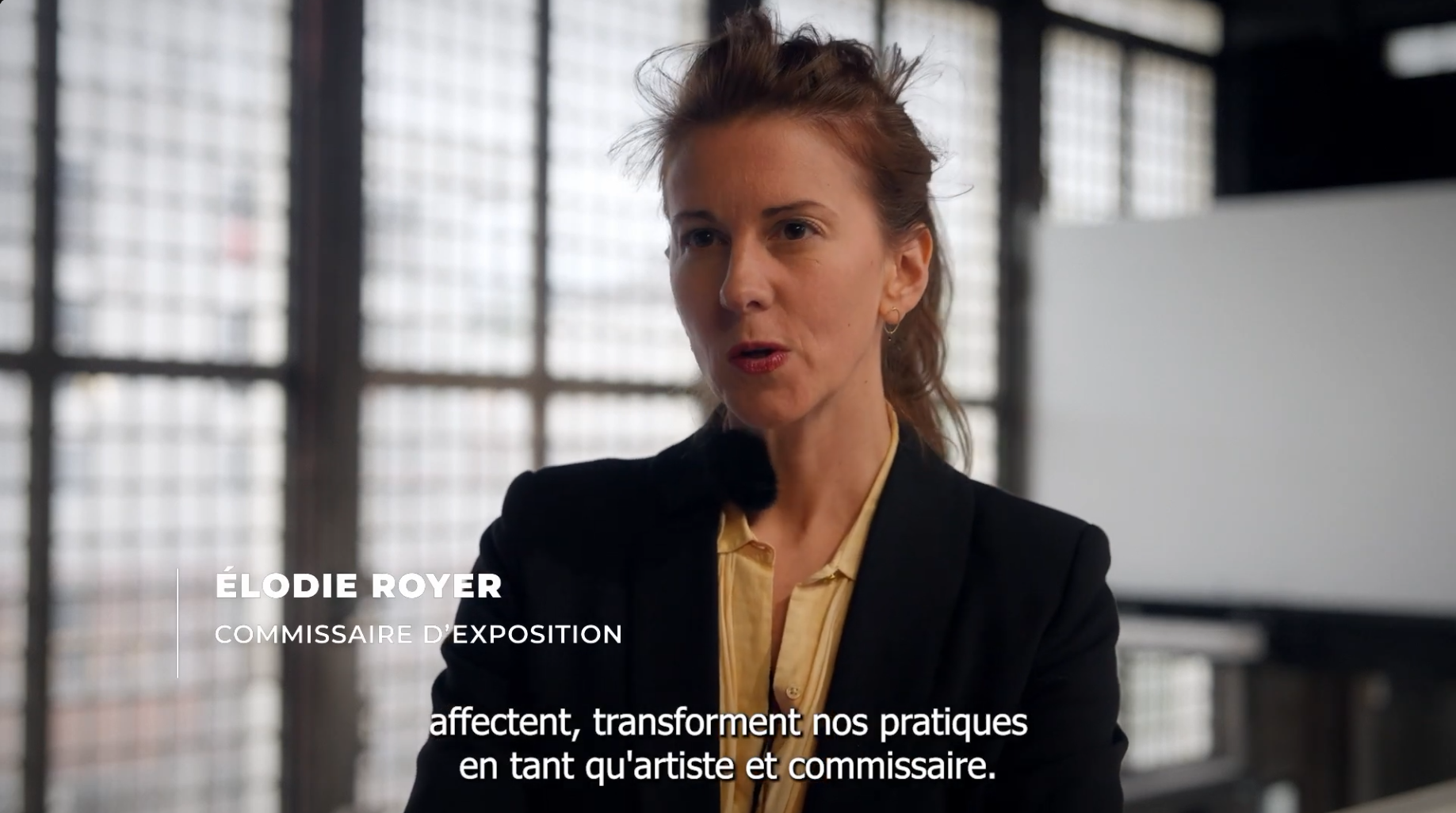 Une femme avec des cheveux bouclés portant une veste noire et une chemise jaune, parlant dans un lieu lumineux avec de grandes fenêtres en arrière-plan.