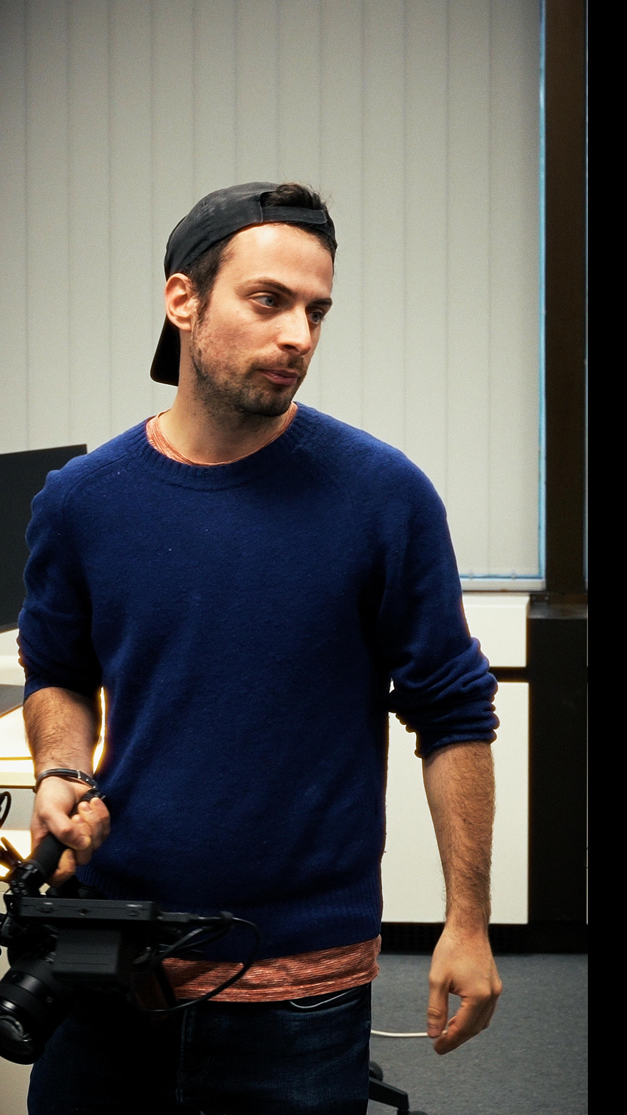 Un homme portant une casquette noire et un pull bleu marine , tient sa caméra, prêt à filmer.