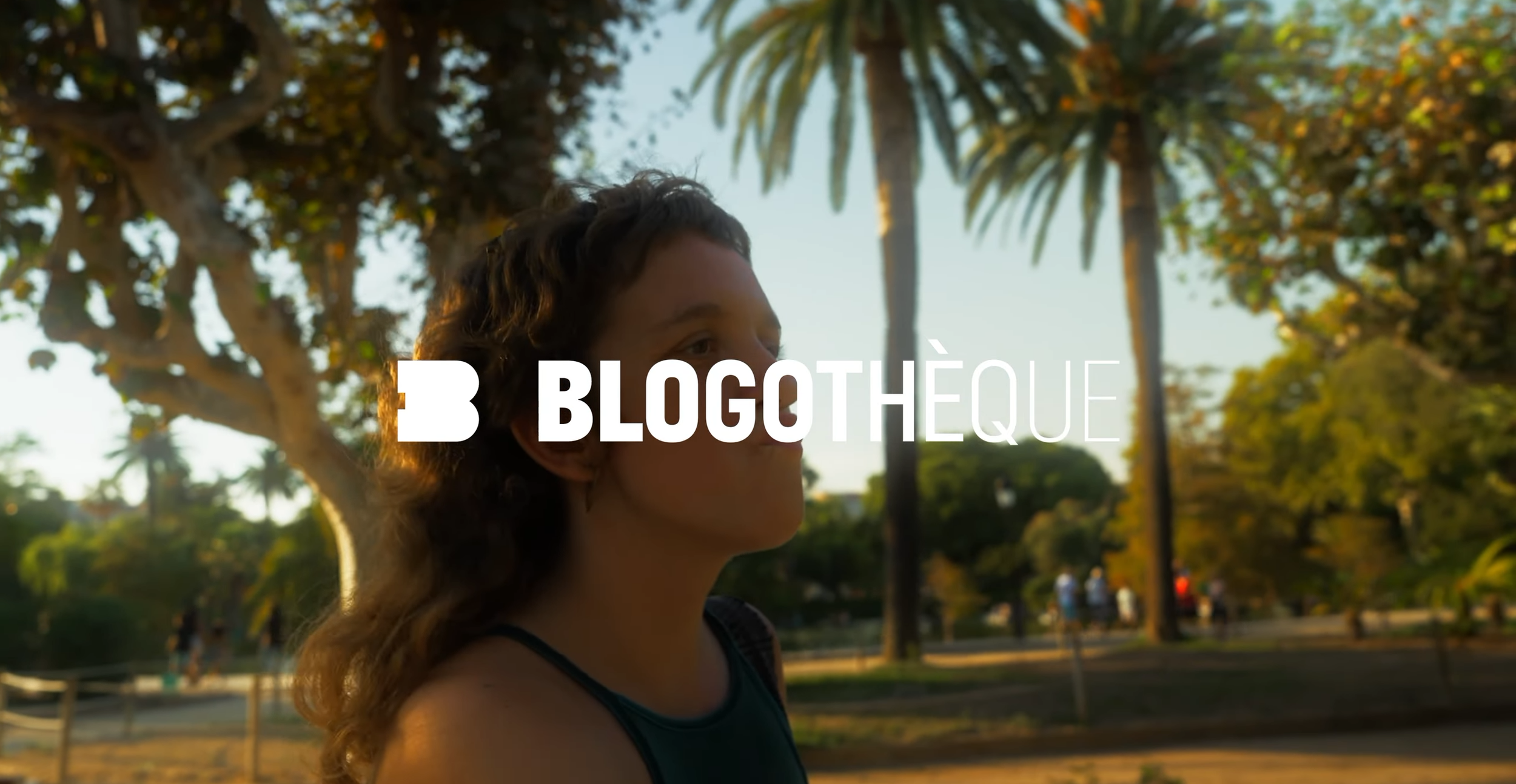 Une femme avec des cheveux bouclés regarde de profil, en arrière-plan un parc avec des palmiers et d'autres personnes.