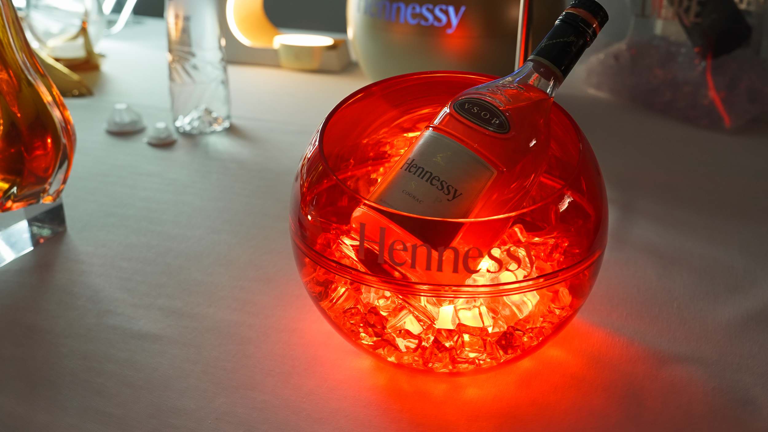 Bouteille de cognac Hennessy dans un seau en verre avec lumière rouge, entourée de glaçons, sur une table plate.
