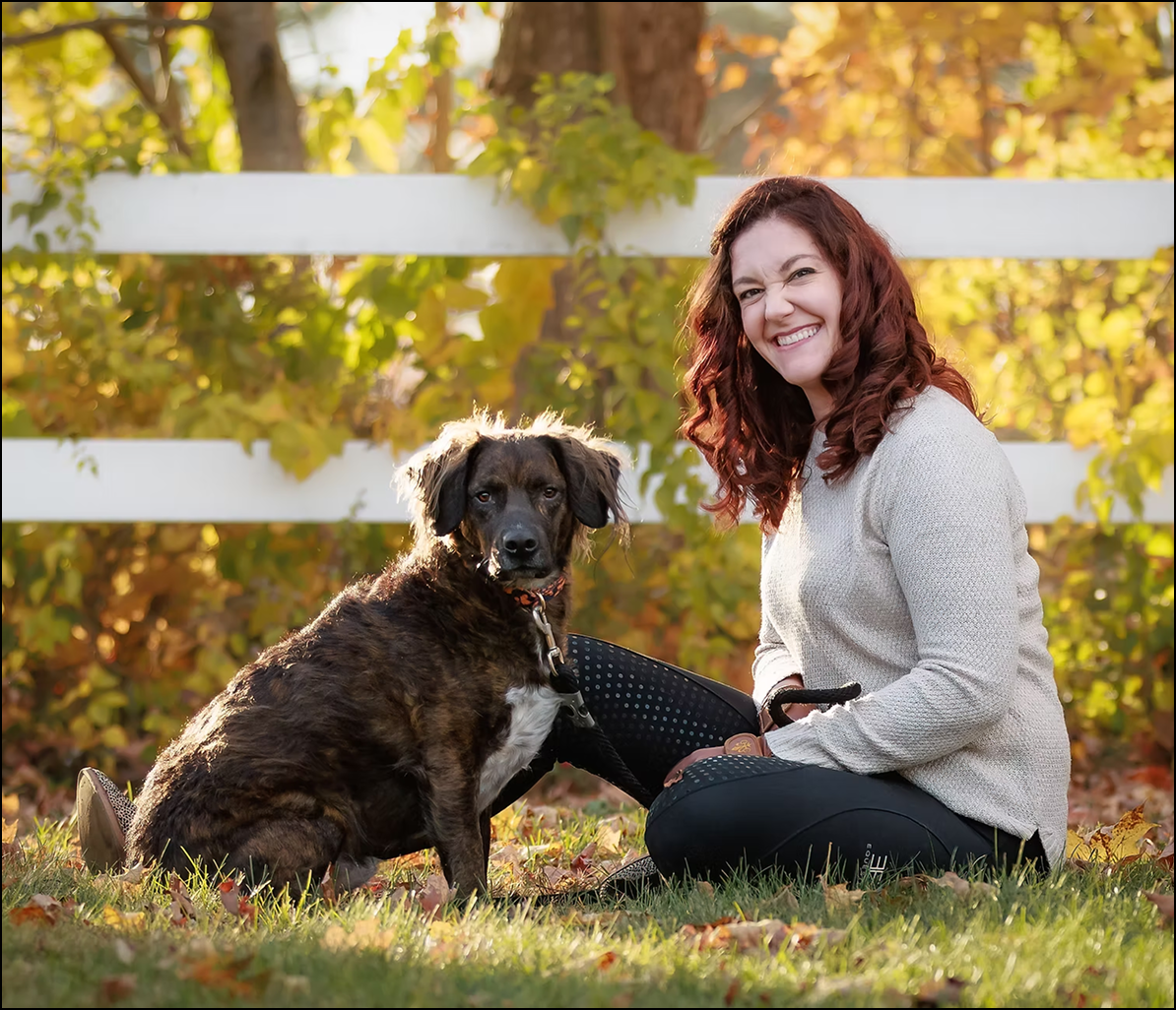 Smiling woman sitting outdoors beside a brown and black dog in an autumn setting