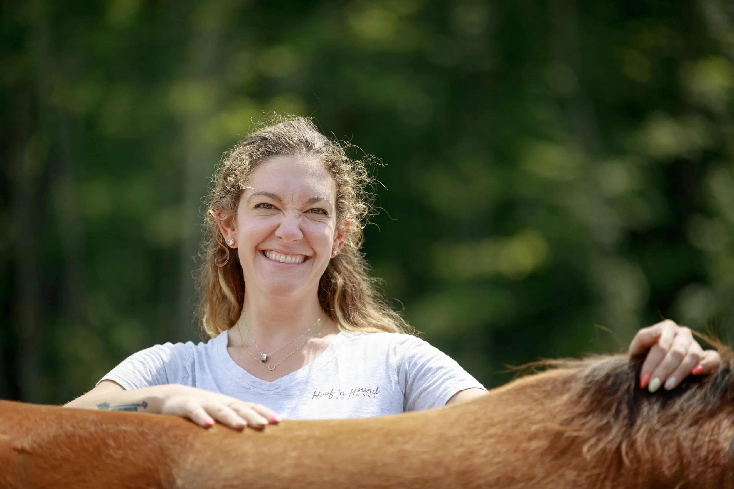 Smiling woman standing outdoors with her hands resting on a brown horse