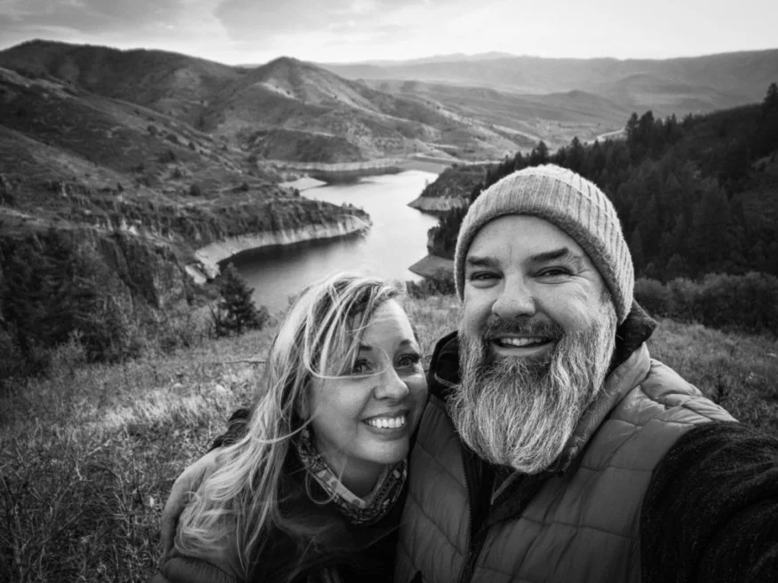 A black and white photo of a smiling couple taking a selfie outdoors with a river and hills in the background.