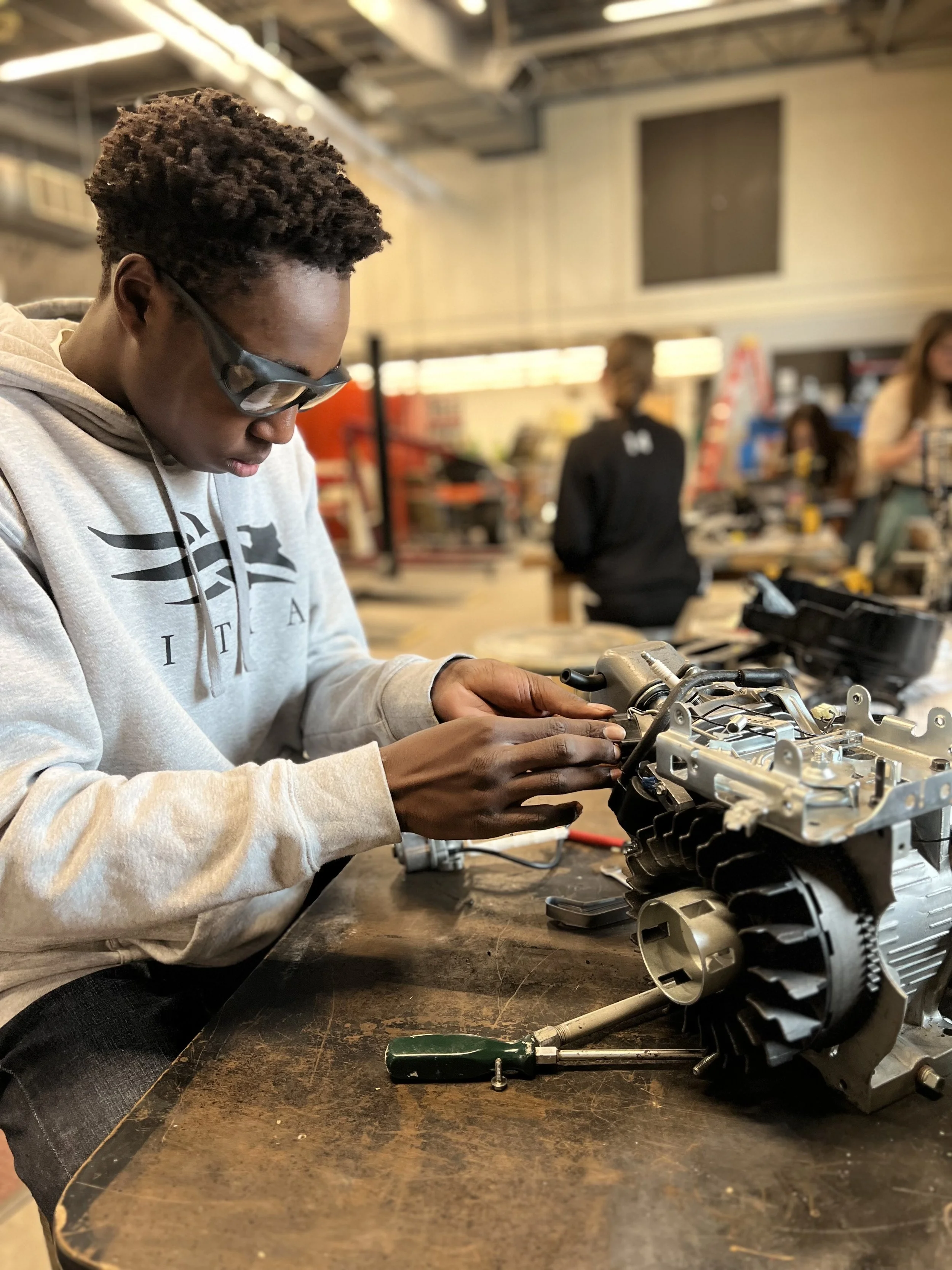 A Fosston high school student wearing glasses and a light-colored hoodie working on an engine or mechanical part at a workshop.