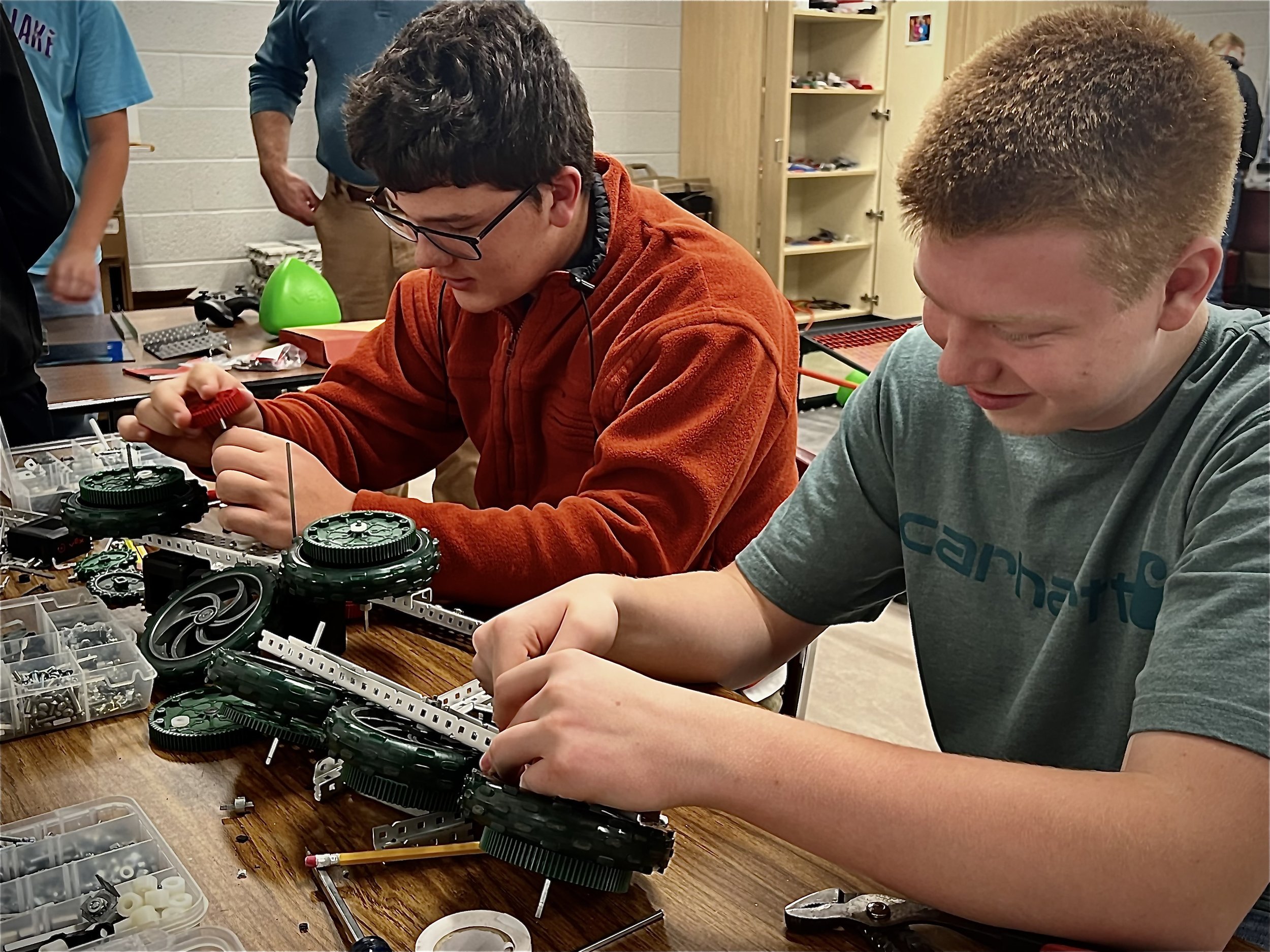 Two students working on assembling a robot in a workshop with shelves and other people in the background.