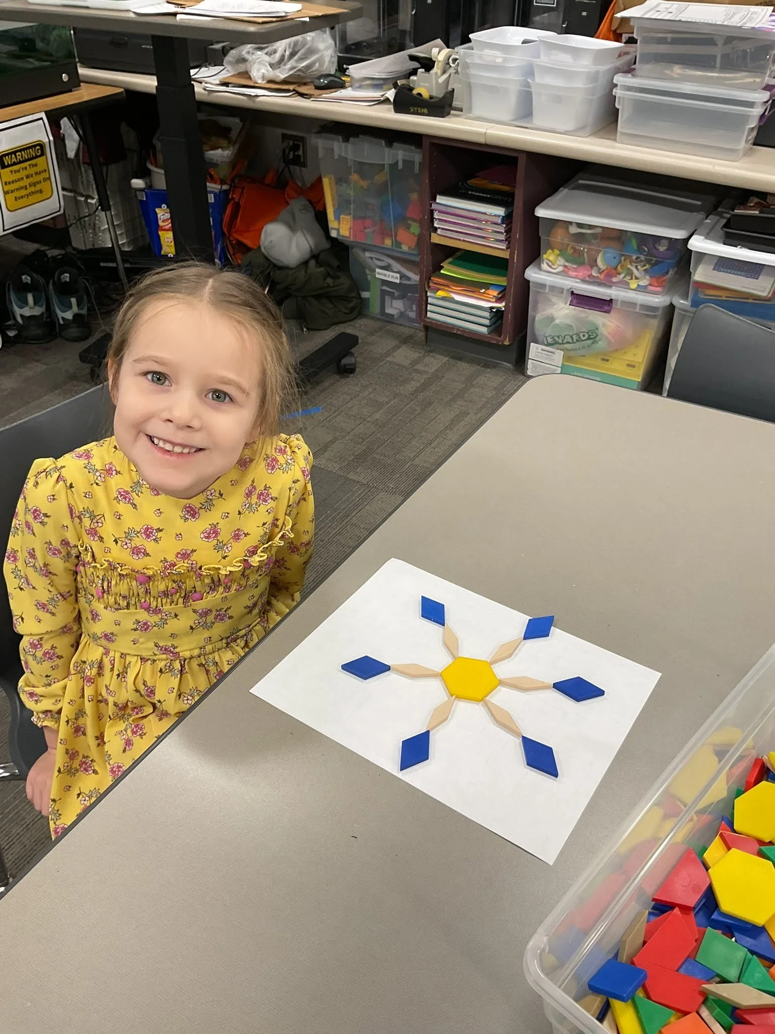 A young girl with a big smile wearing a yellow floral dress is seated at a table with a geometric shape made of colored wooden pieces. The background shows shelves filled with plastic containers, books, art supplies, and miscellaneous items.