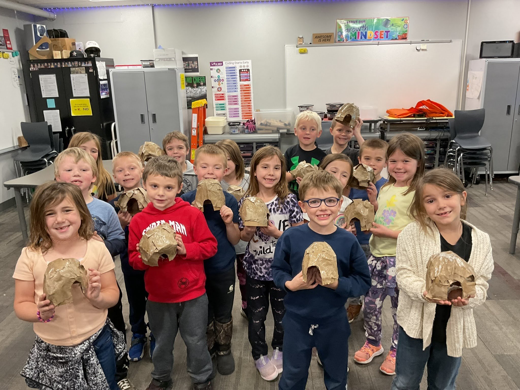 A group of children standing in a classroom, holding paper-mâché skulls. They are smiling and appear to be participating in an art activity.