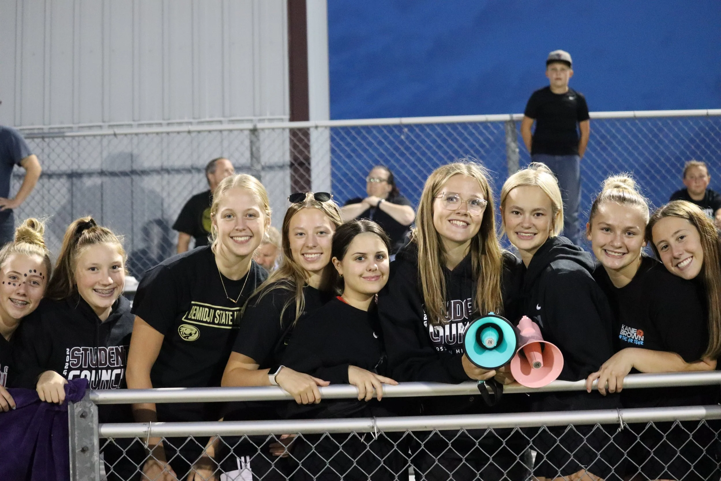 Group of smiling young students gathering at a sports event behind a chain-link fence, with people and a blue sky in the background.