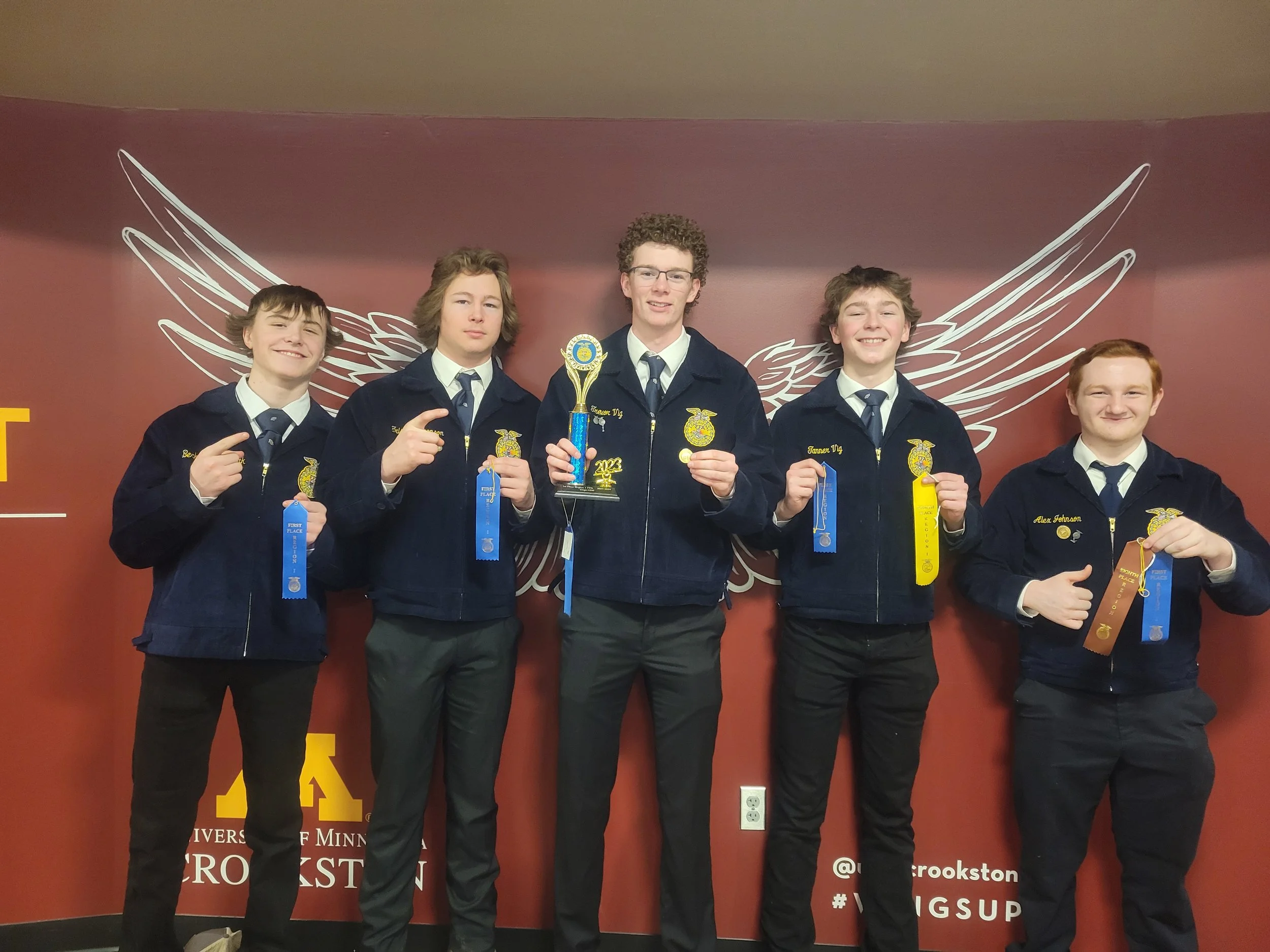 Five young men in matching dark jackets and ties standing in front of a maroon wall with a winged emblem. The middle man holds a trophy and blue ribbon, while the others display various ribbons and medals, smiling and pointing.
