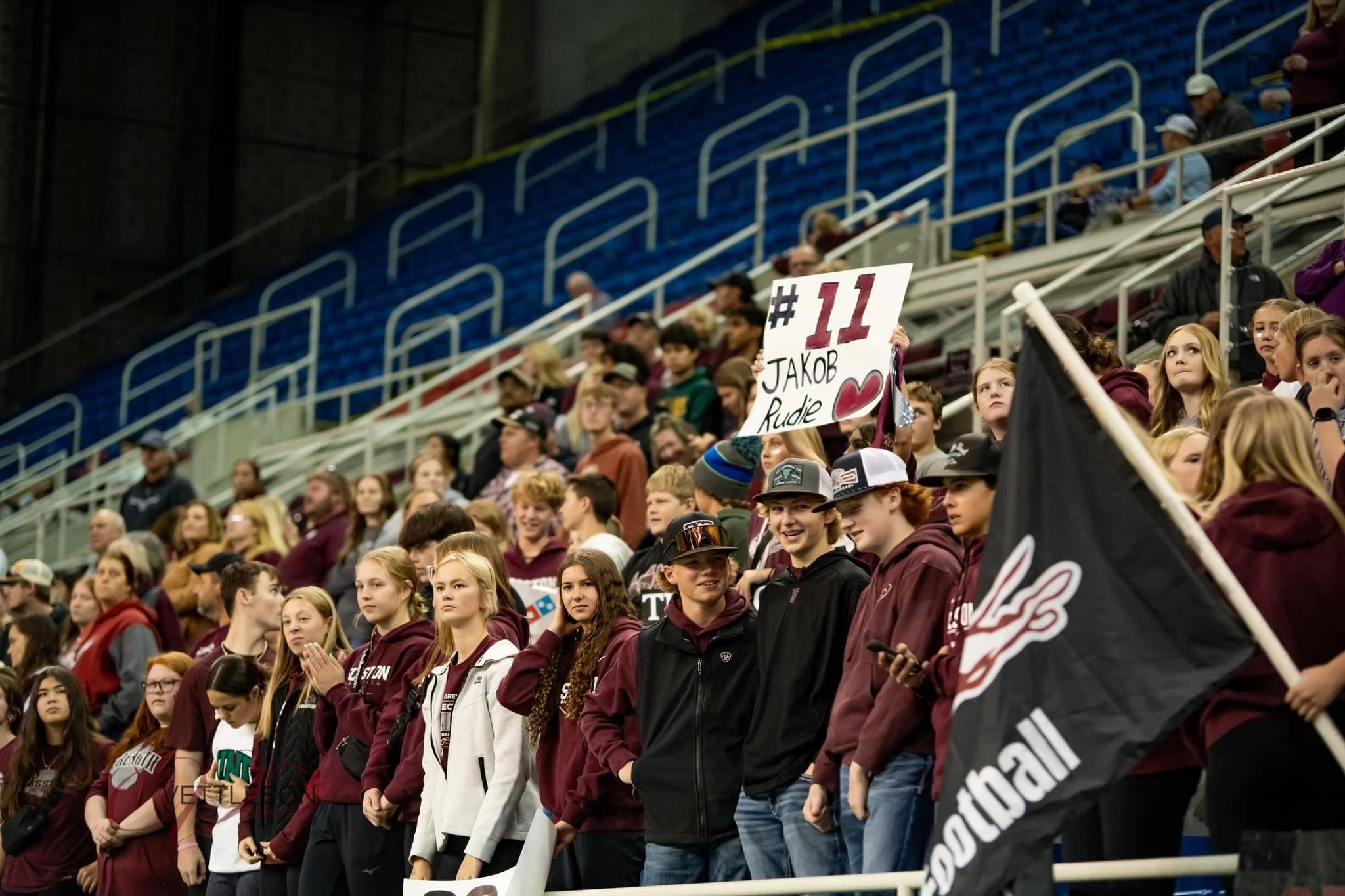 A crowd of Fosston Public Schools students standing in stadium seats at a high school sports game, some holding signs, wearing maroon and white clothing.