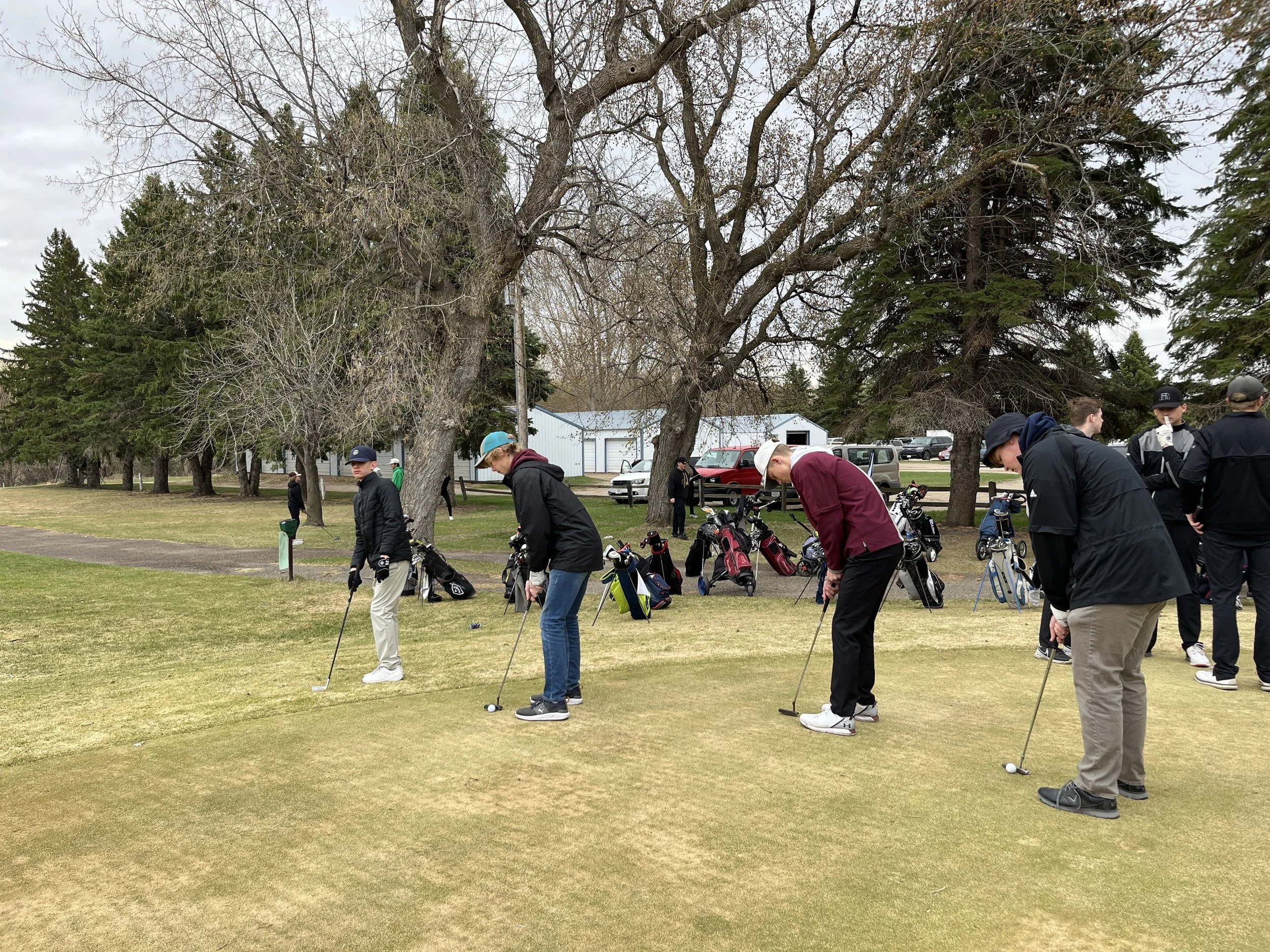 A group of students playing golf on a grassy course surrounded by trees, with golf equipment and a building in the background.