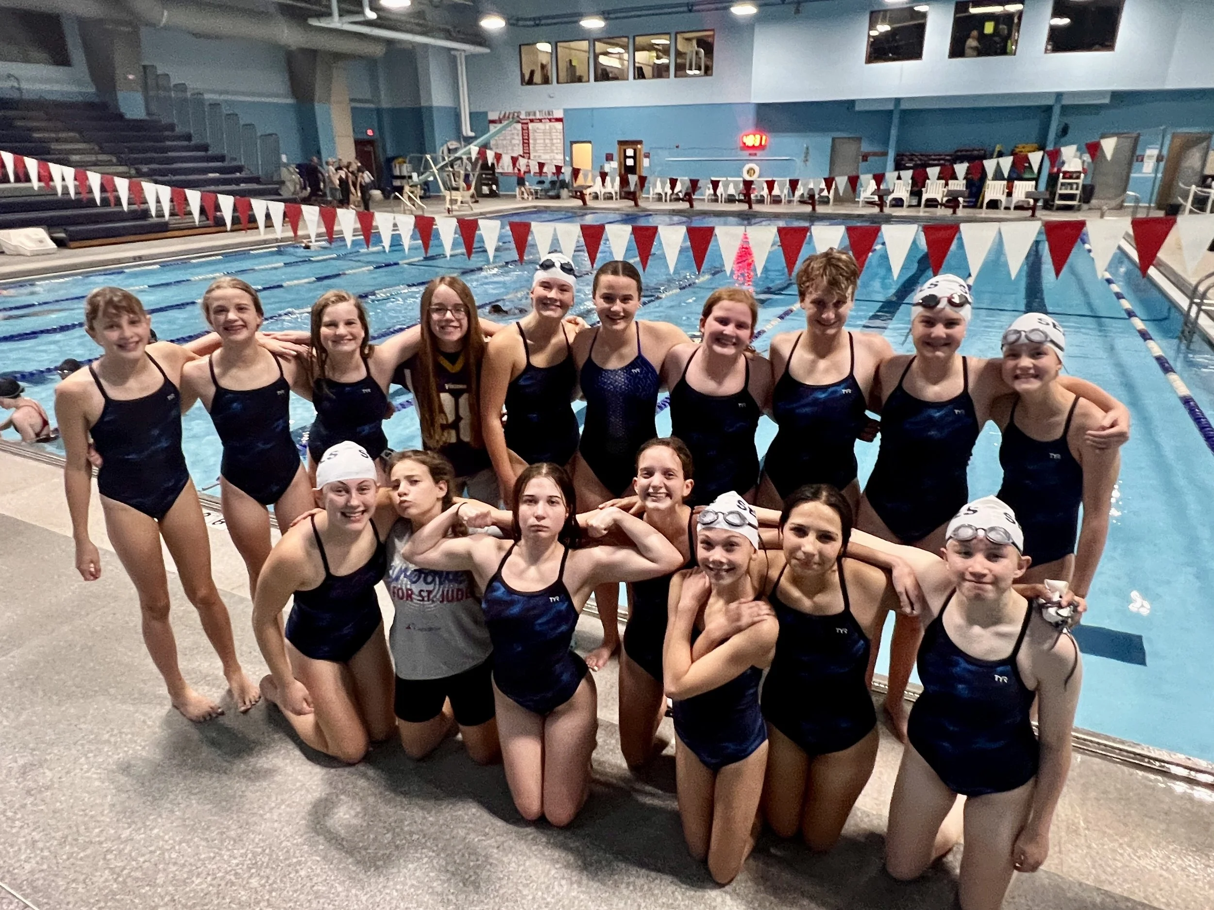A group of students on the Fosston Girls swim team in black swim caps and swimsuits, posing together at an indoor swimming pool after a swim meet. They are smiling and standing close to each other in front of the pool.