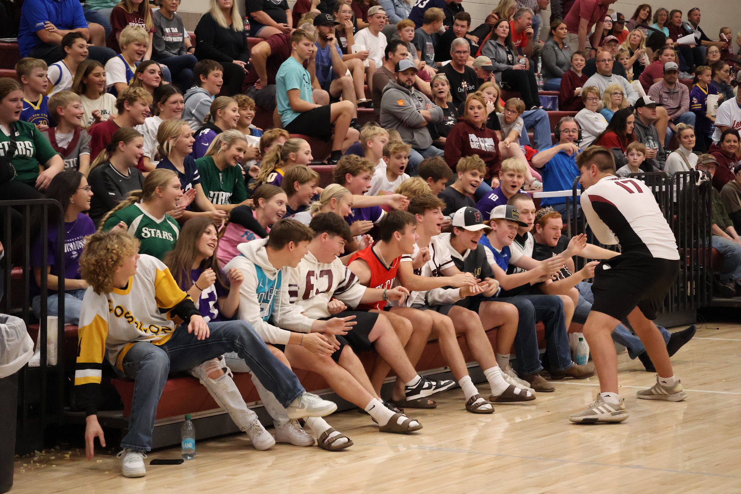 Crowd of young kids and teenagers sitting in stadium seats, watching and reacting with excitement and laughter, at an indoor sports event.