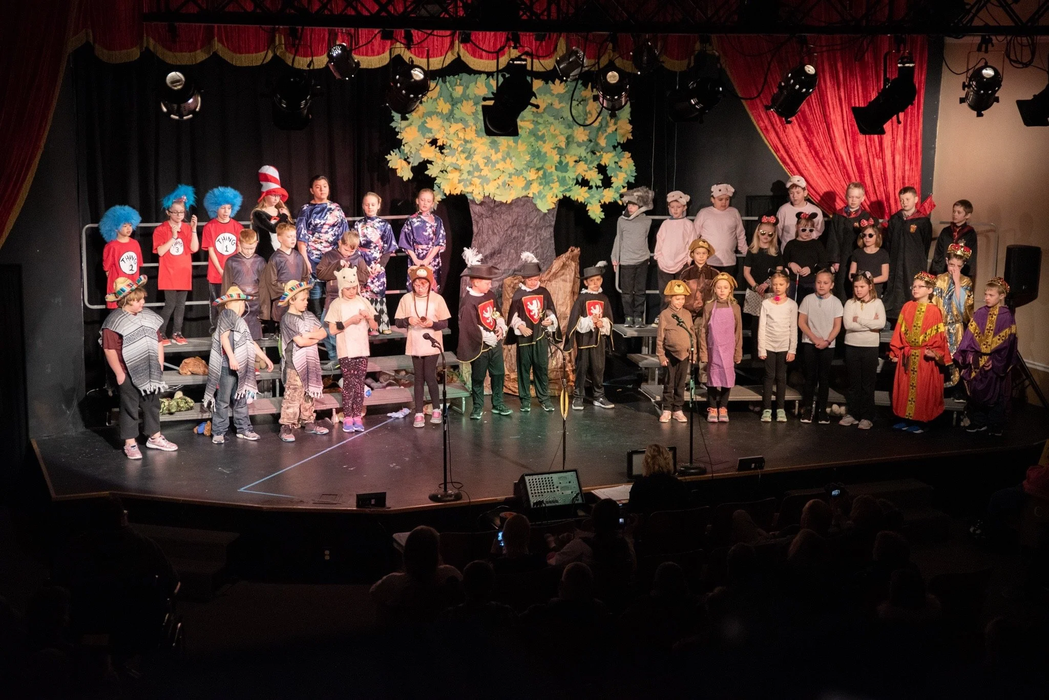 Children performing on stage during a school play, dressed in various costumes including animals, pirates, and characters from fairy tales, with a backdrop featuring a large tree and yellow leaves.