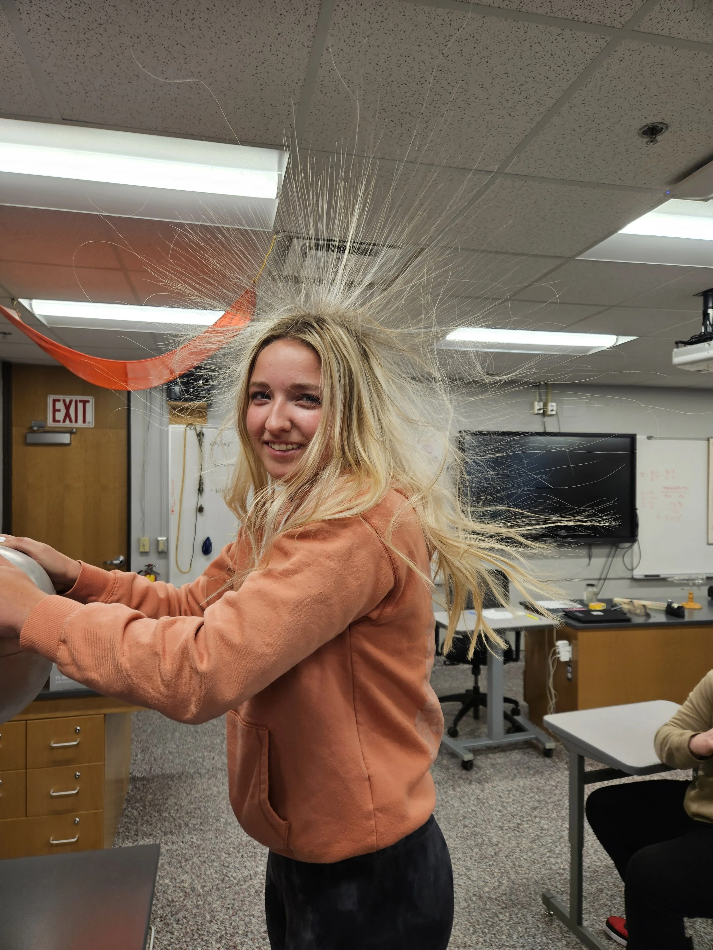 Young student with long blonde hair in a classroom, mid-laugh, hair splaying outward as if caught in a gust of wind, wearing an orange hoodie, with classroom equipment in the background.