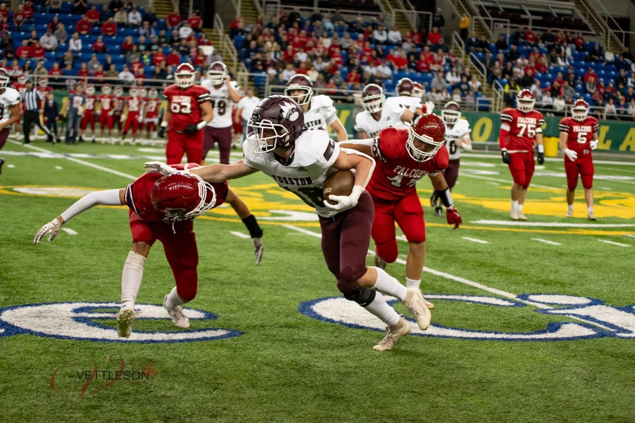 A high school football game in progress. A player in a white jersey with black helmet is running with the ball, while players in red jerseys are trying to tackle him. Spectators are seated in the stands in the background.
