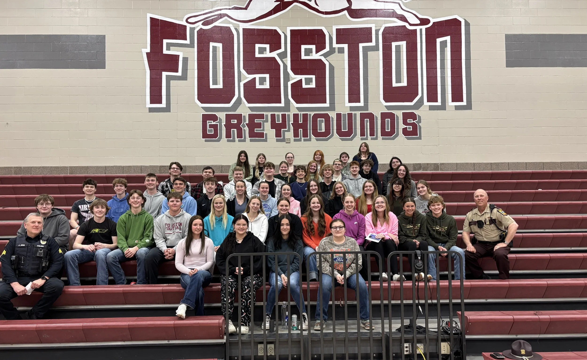 A large group of students and two adults, one in a police uniform, posing for a group photo in a gymnasium with a maroon and gray bench seating. Behind them is a large wall mural that reads 'PERSISTENCE GREYHOUNDS' with a stylized graphic above the t