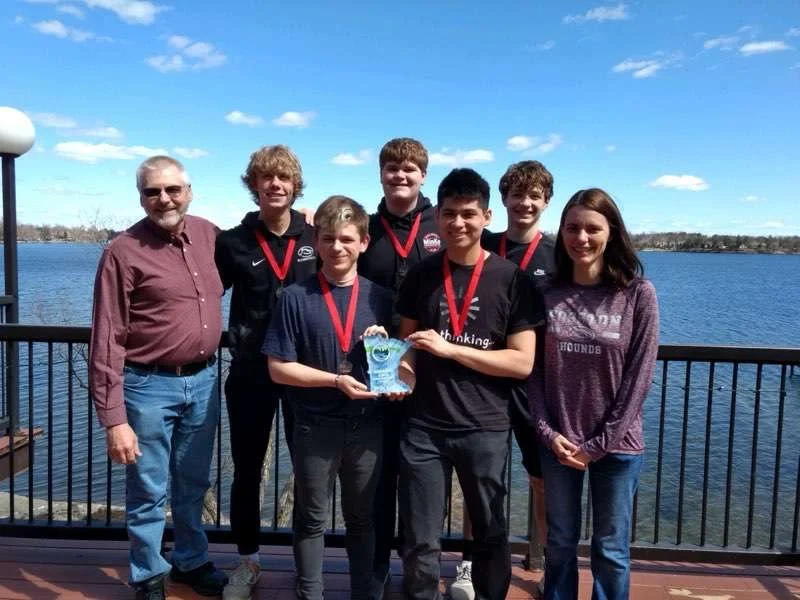 Group of six students and one adult standing on a pier by the water, holding a trophy and wearing medals, with a lake and blue sky in the background.