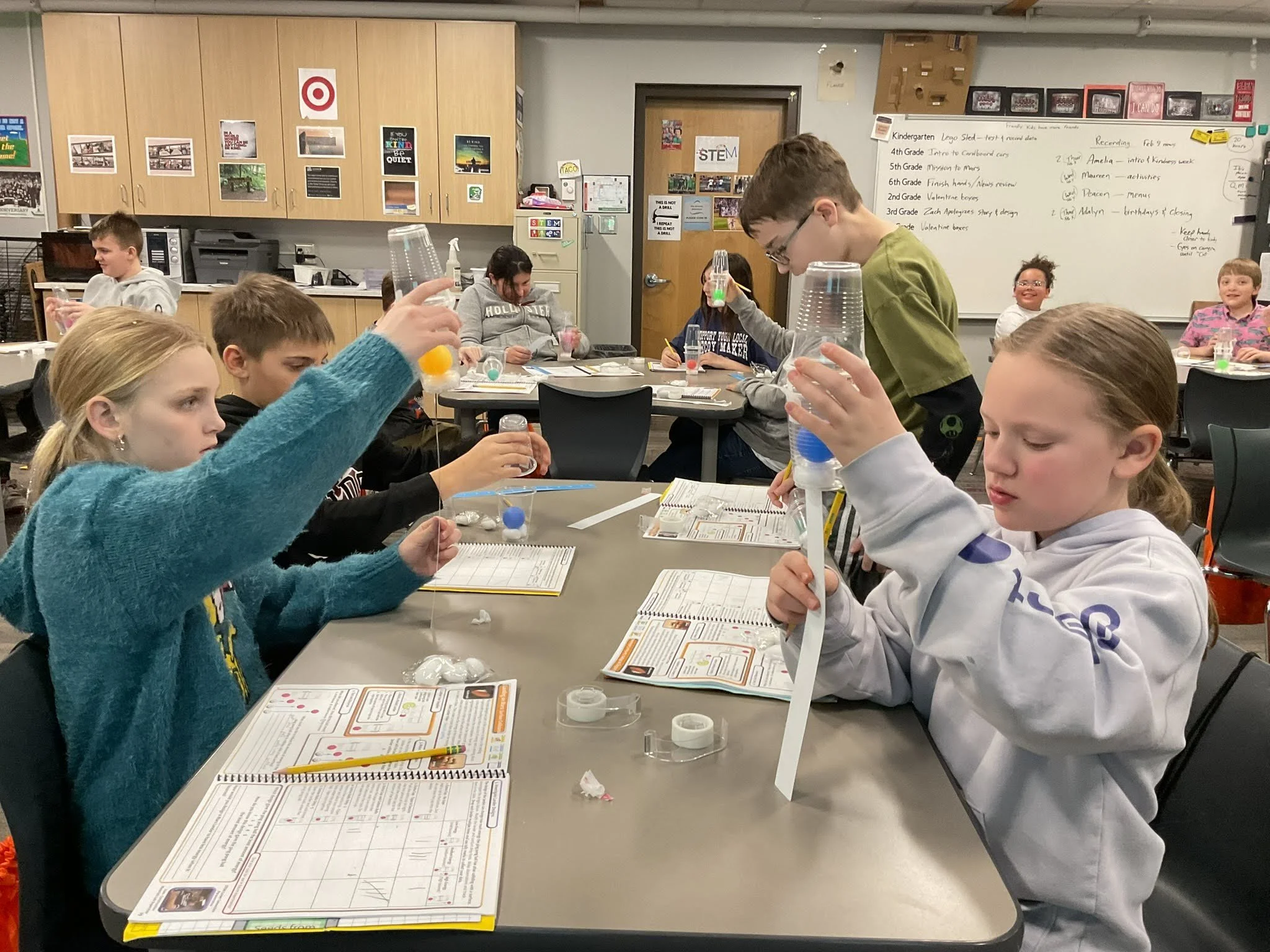 Children in a classroom sitting at tables engaged in a science activity with various science supplies, notebooks, and worksheets, with a teacher assisting them.