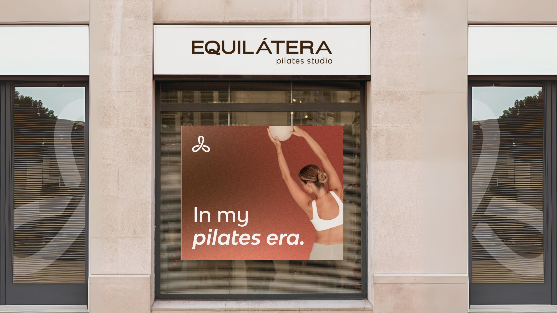 Cartel en una ventana de un estudio de pilates llamado EQUILÁTERA, con una mujer haciendo ejercicio con una pelota, y un texto que dice "In my pilates era".