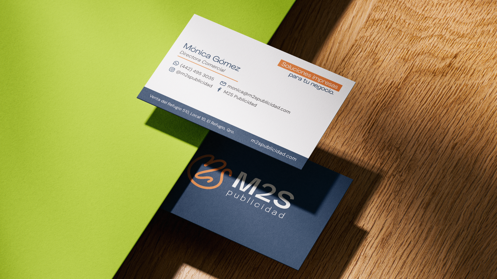 Tarjetas de presentación de una empresa de publicidad, una en enfoque y otra parcialmente visible, sobre una mesa de madera y fondo color verde y negro.