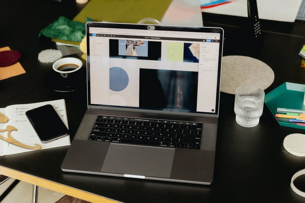 Computadora portátil abierta en una mesa de trabajo con una pantalla con aplicaciones abiertas, junto a una taza de café, un teléfono móvil, un vaso de agua y varios objetos de oficina en el entorno.