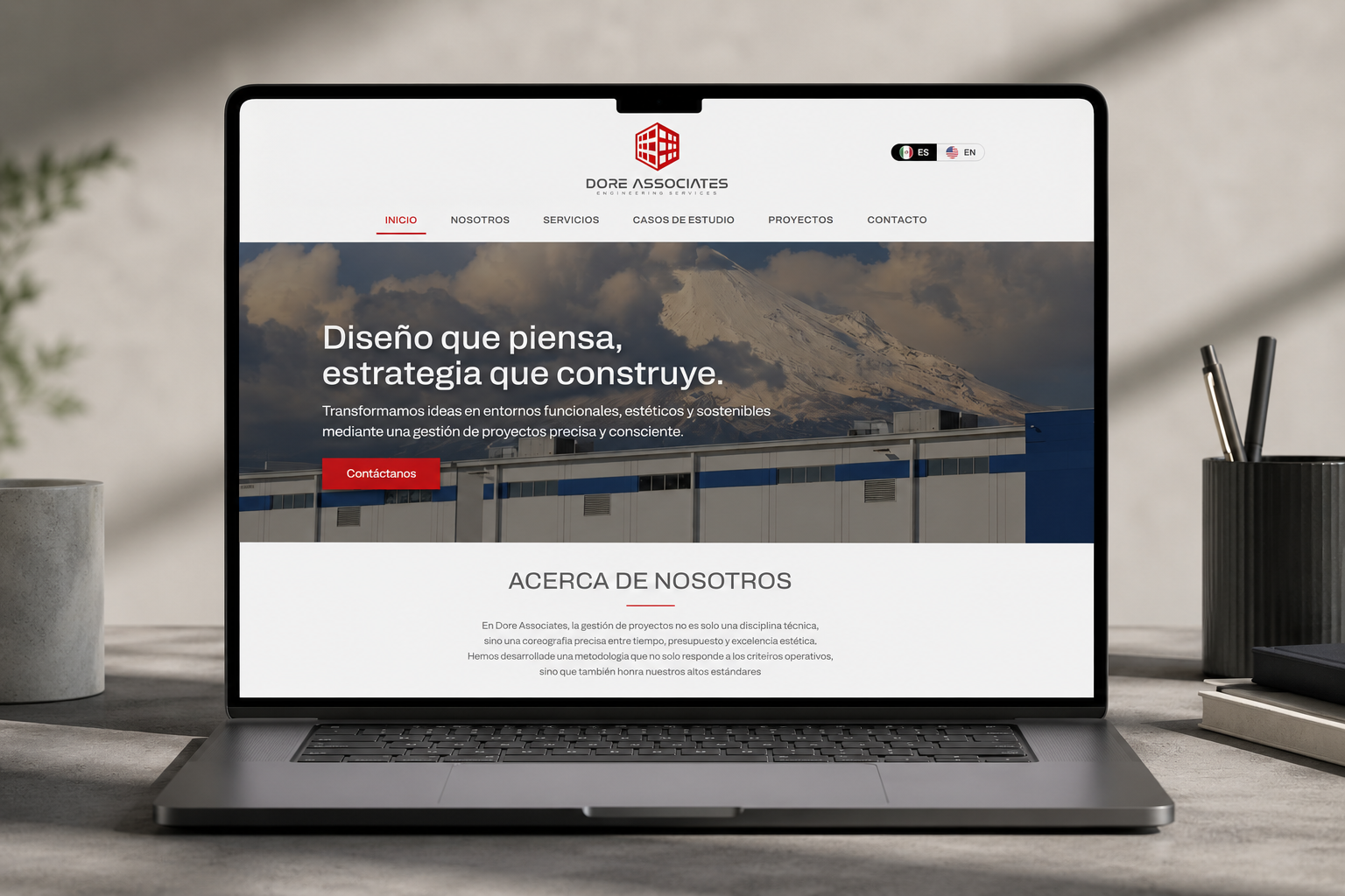 Pantalla de laptop mostrando la página web de Dore Associates, empresa de servicios de ingeniería, con un fondo de montañas y edificios, en un escritorio con bolígrafos y libros