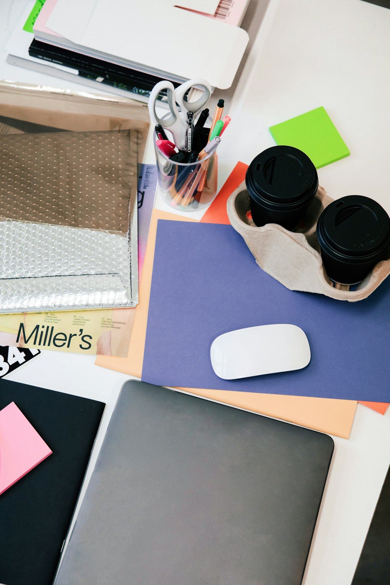 Mesa de oficina con papelería, tijeras, cafés para llevar, blocs de notas de colores, mouse y computadora portátil.