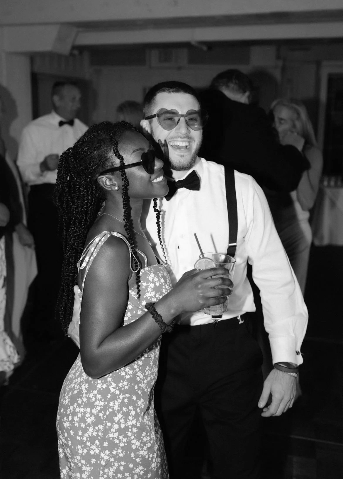Black and white photo of a young woman and man dressed in formal attire at a party, smiling and holding drinks. The woman is wearing a floral dress and glasses, the man is wearing a white shirt, bow tie, and suspenders. Other people are seen in the background, some dancing.