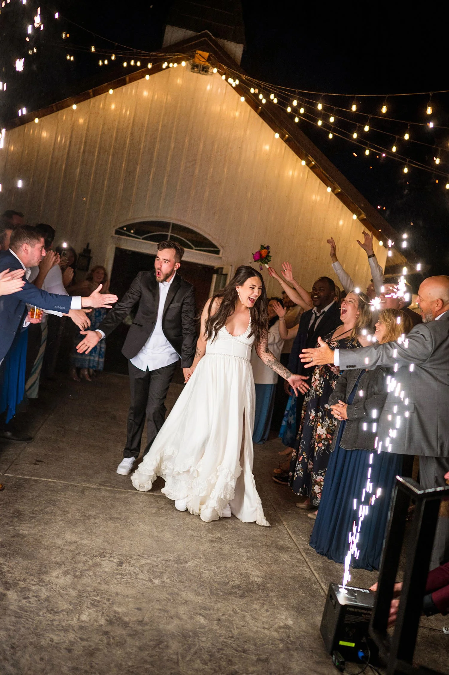 Bride and groom dancing at their wedding reception surrounded by guests, with string lights hanging from the ceiling and sparklers on the floor.
